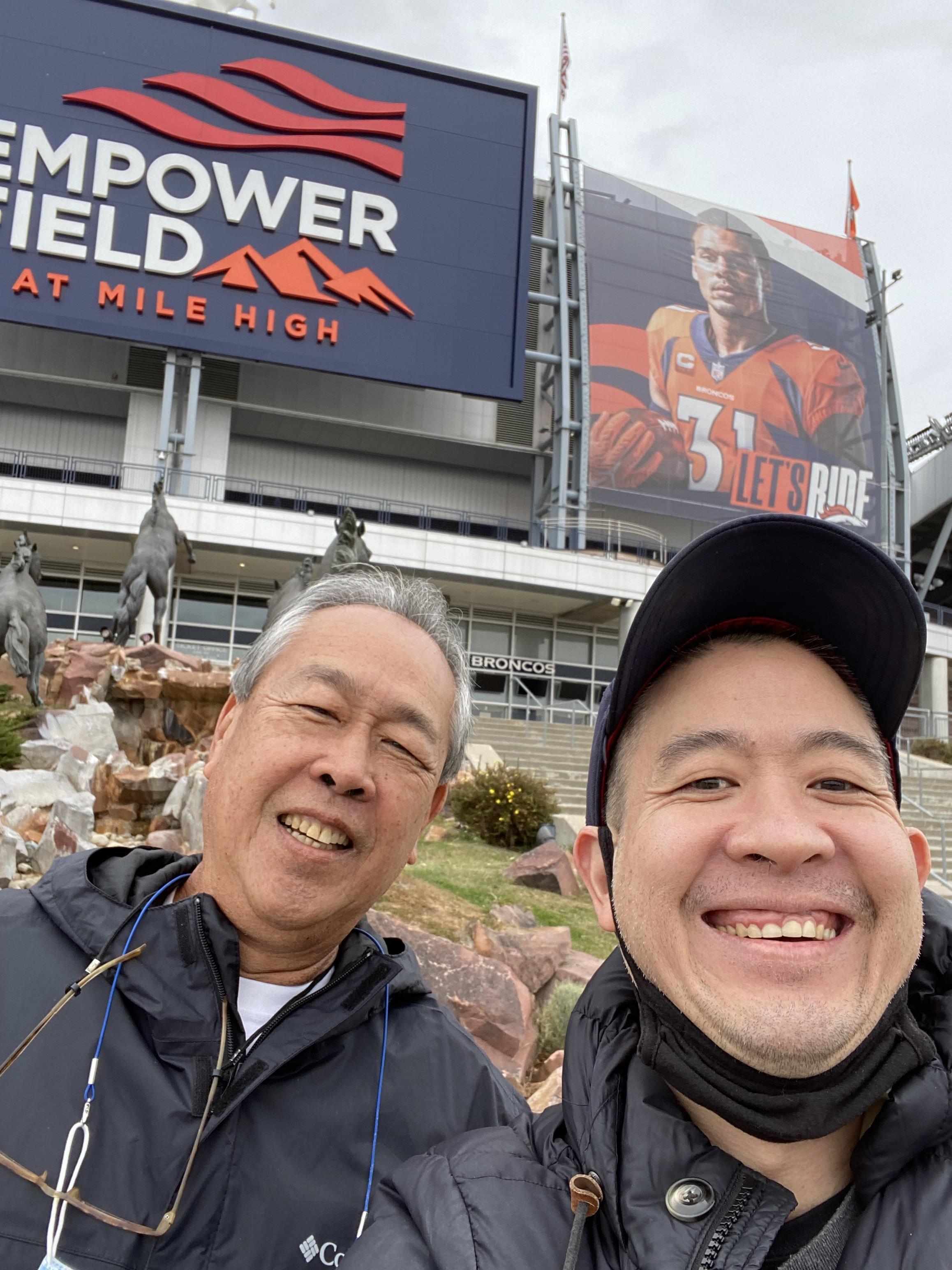 Friends smile together in front of Empower Field, capturing a joyful moment in sports culture.