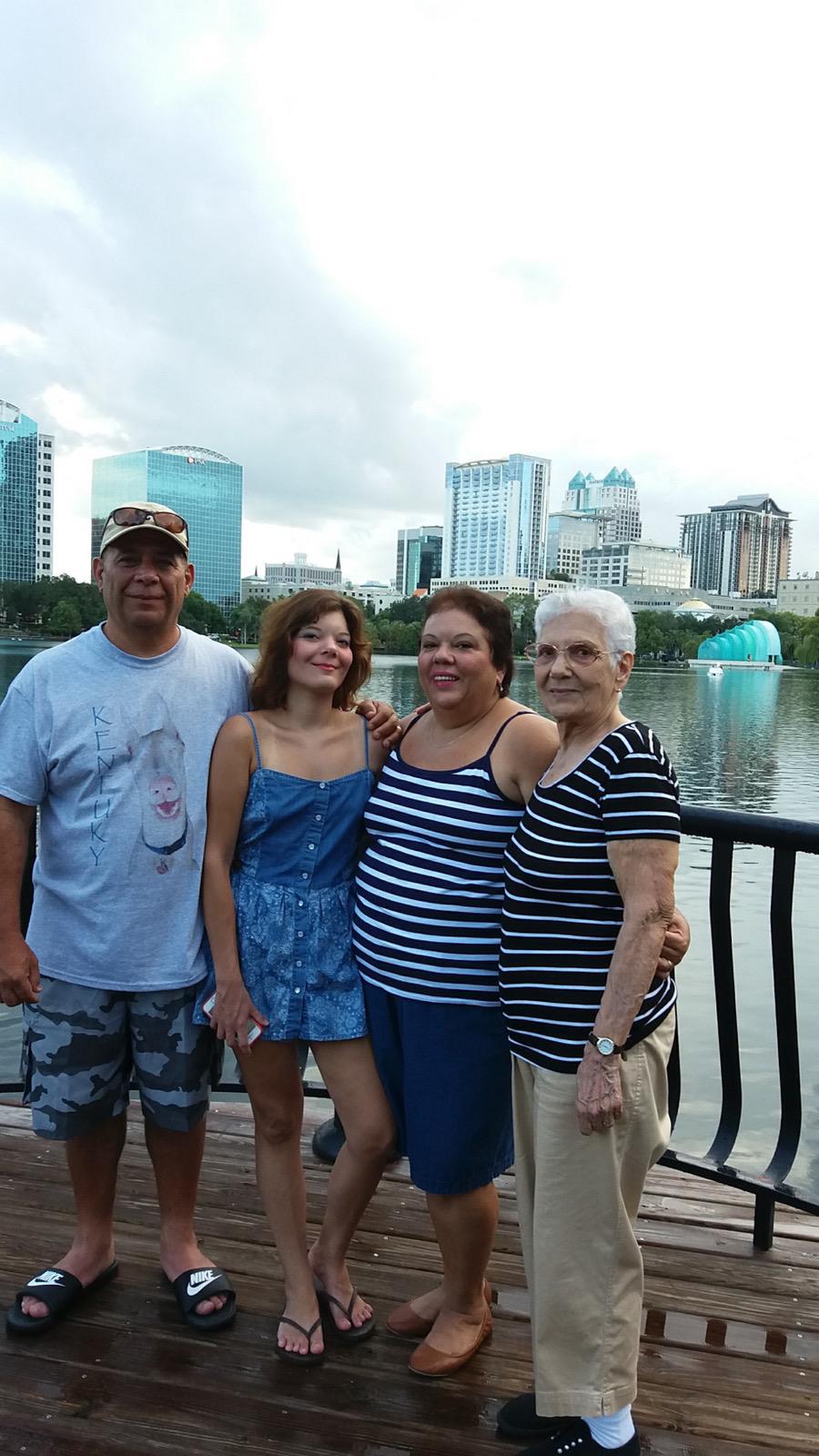 Four family members pose happily together by the lake with city buildings in the background.