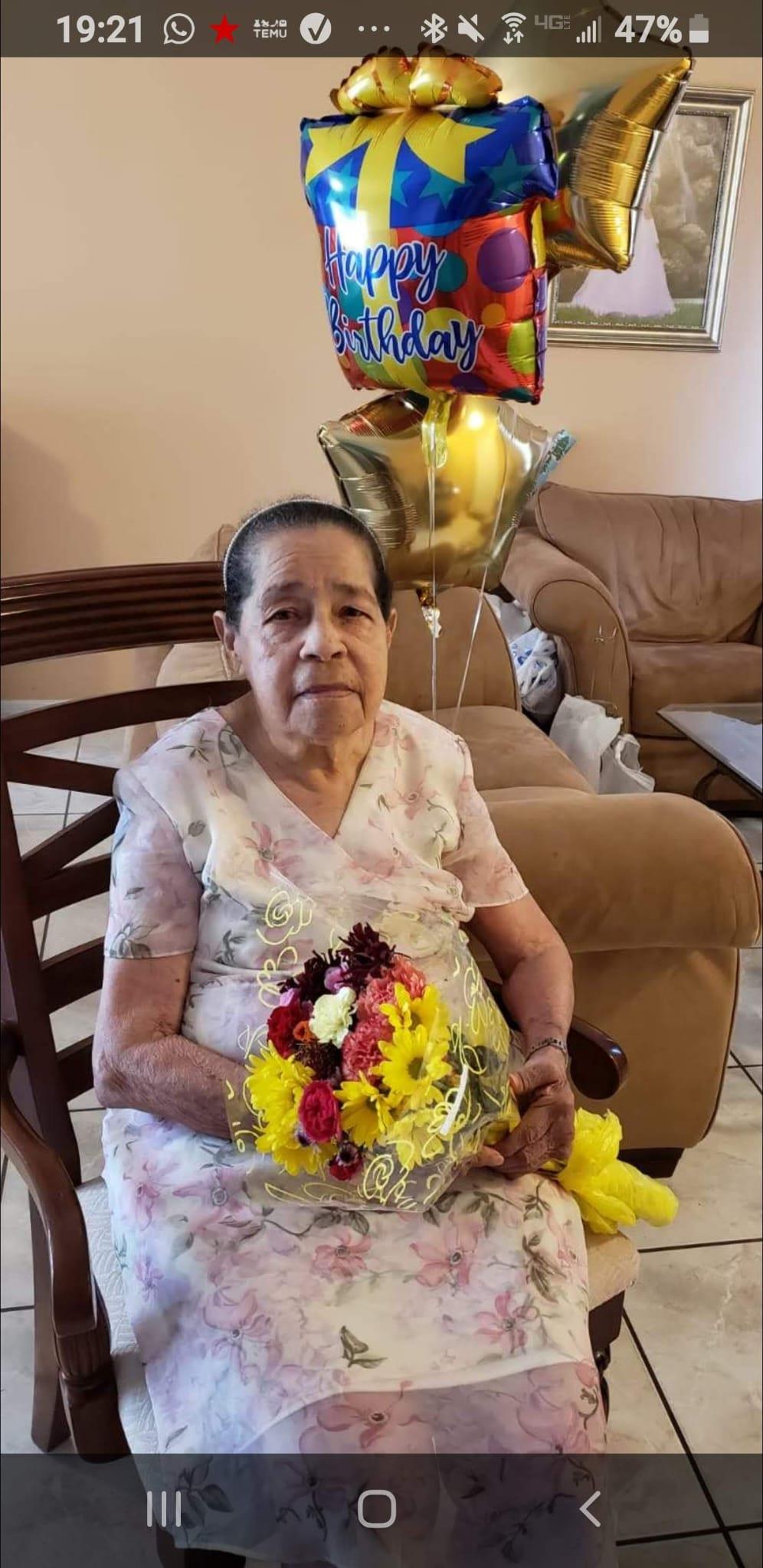 Elderly woman sits on a chair, smiling while holding a bouquet of flowers, celebrating her birthday.