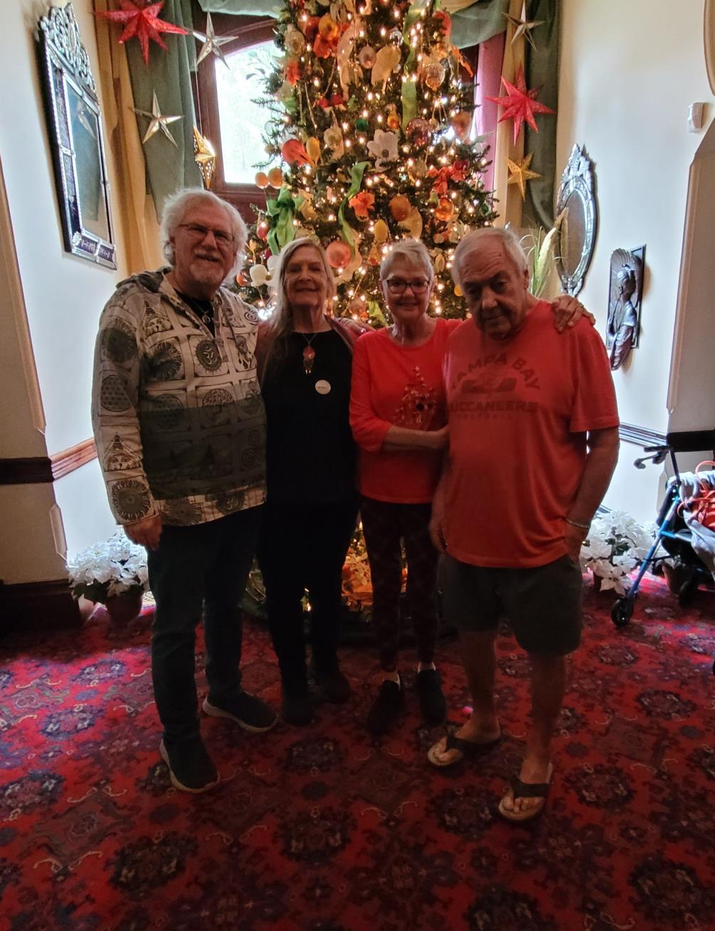 Four friends pose joyfully in festive attire near a stunning Christmas tree adorned with ornaments.