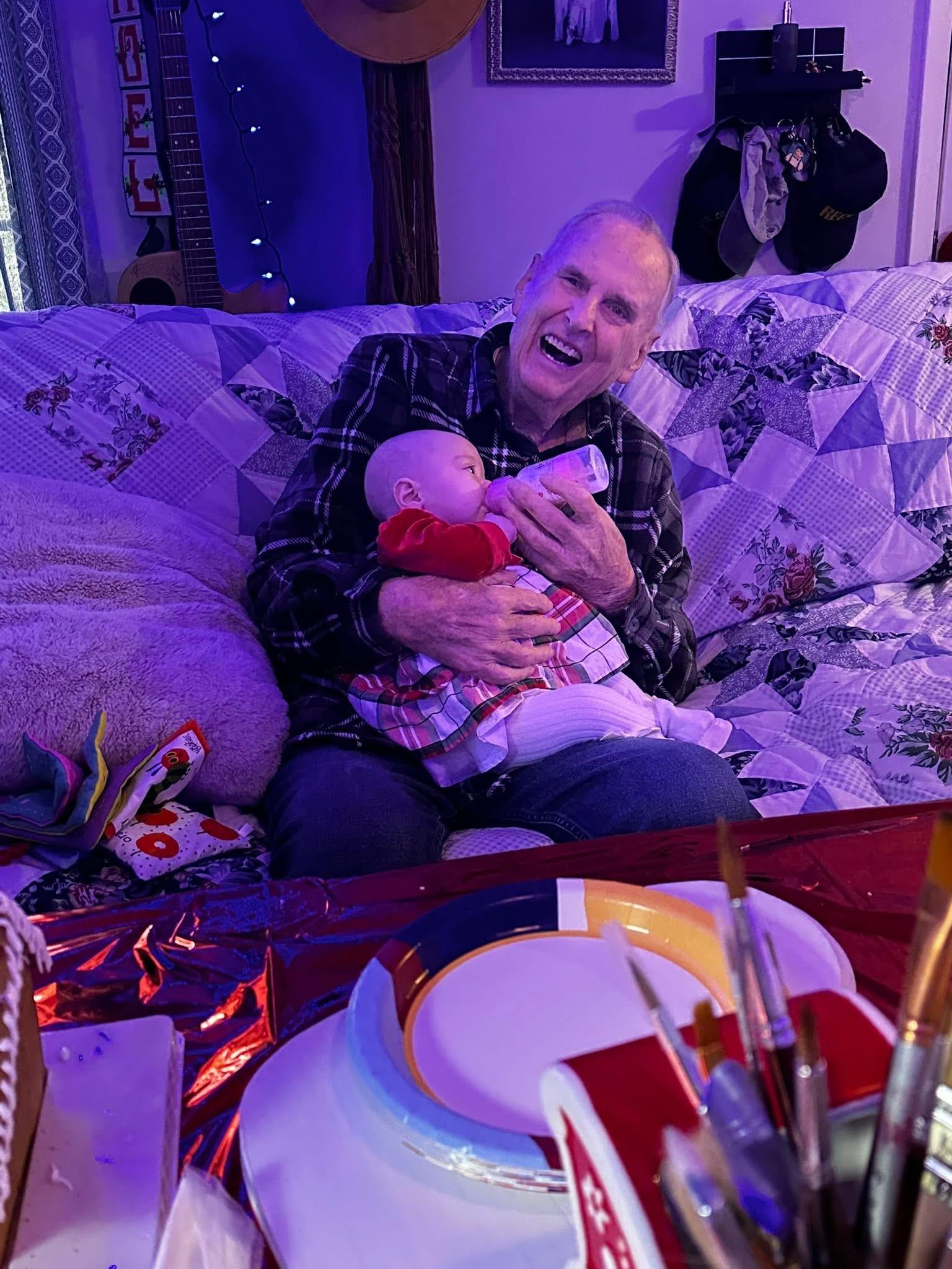 A joyful elderly man cradles an infant in his arms on a cozy couch at a festive indoor gathering.