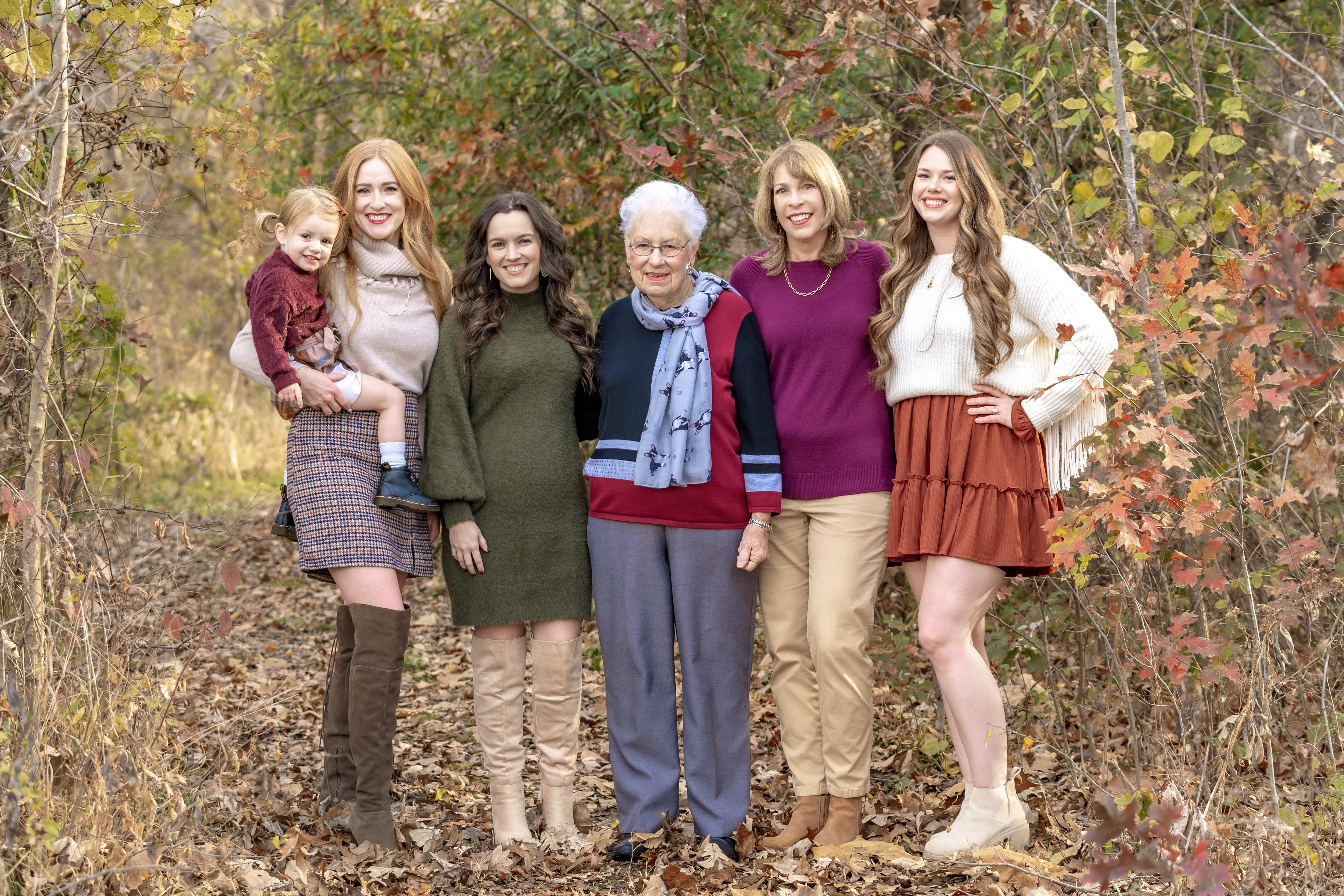 Six women of different ages stand together in a wooded area adorned with autumn foliage.