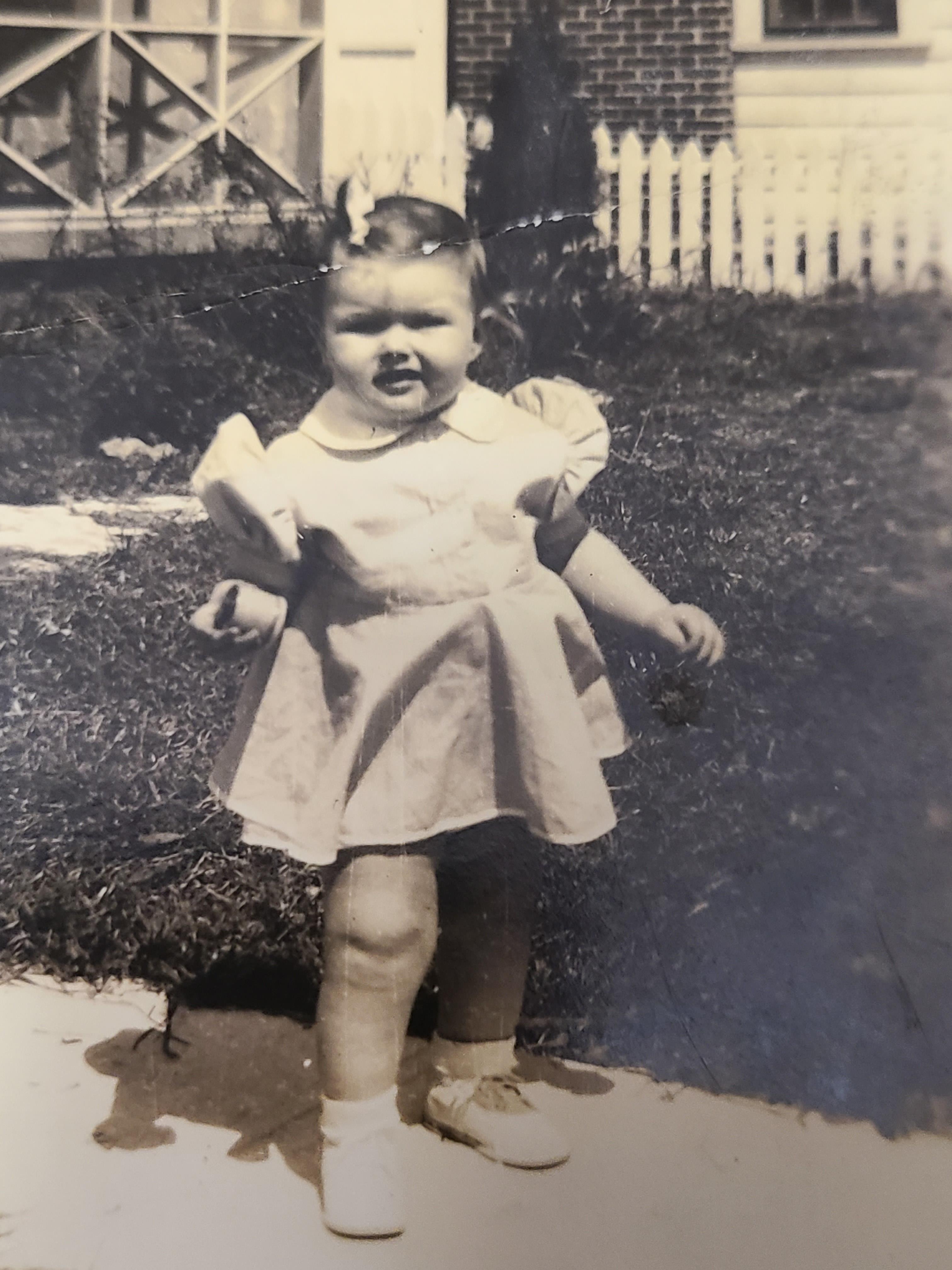 A girl in a vintage dress smiles on a suburban path while holding a snack.
