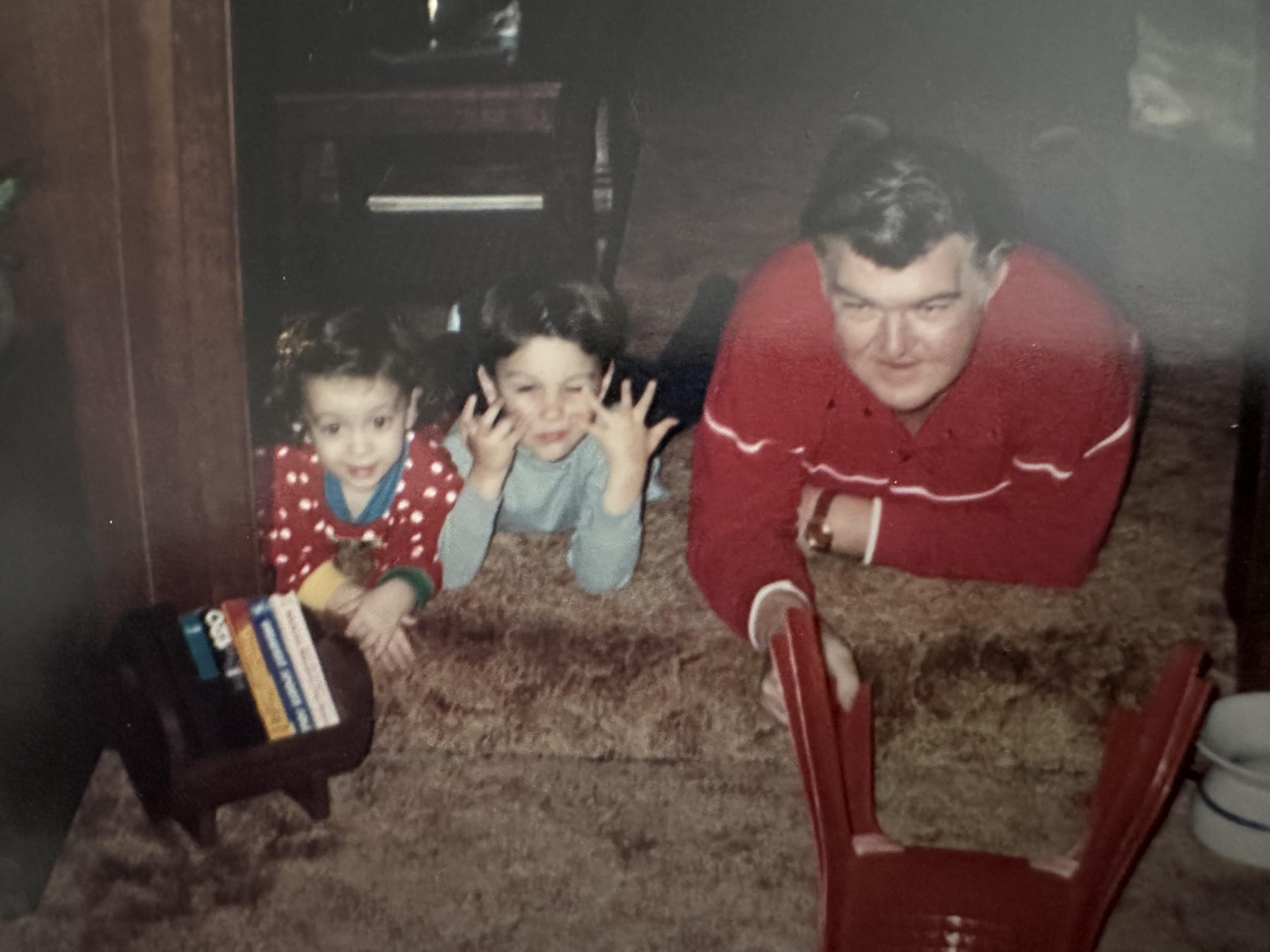 Two children playfully pose with their grandfather on a carpet in a nostalgic living room.