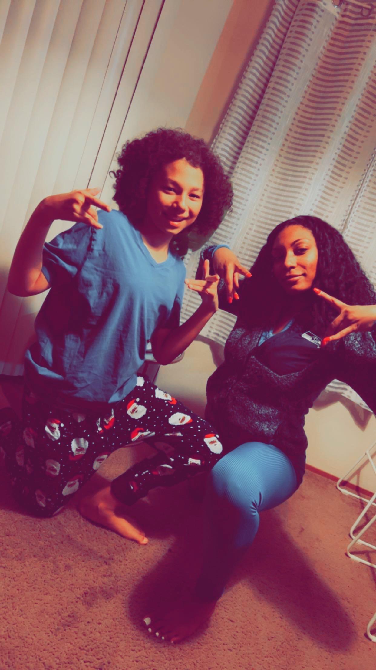 Two friends are crouching down, smiling, and making peace signs while wearing loungewear at home.