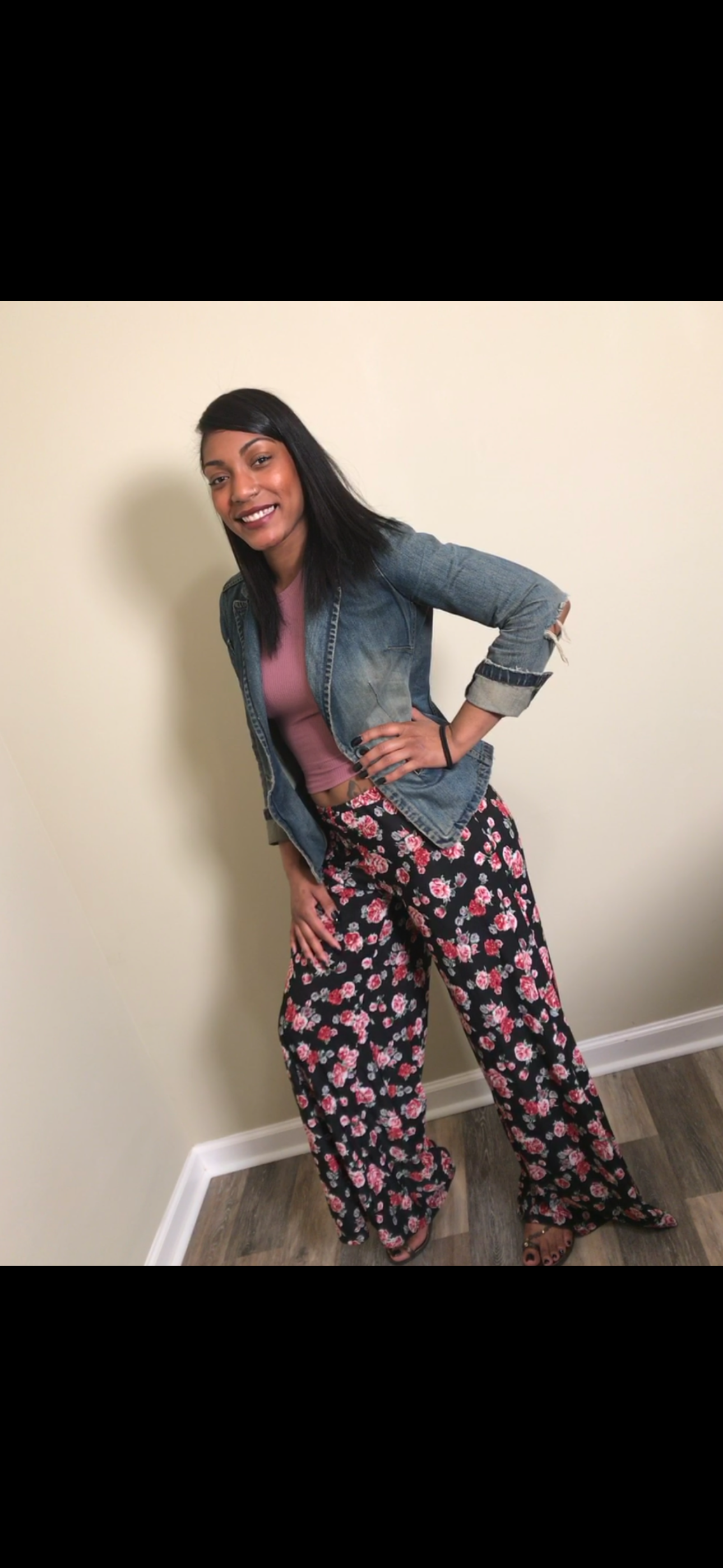 A young woman poses happily, showcasing her floral pants and denim jacket at home.