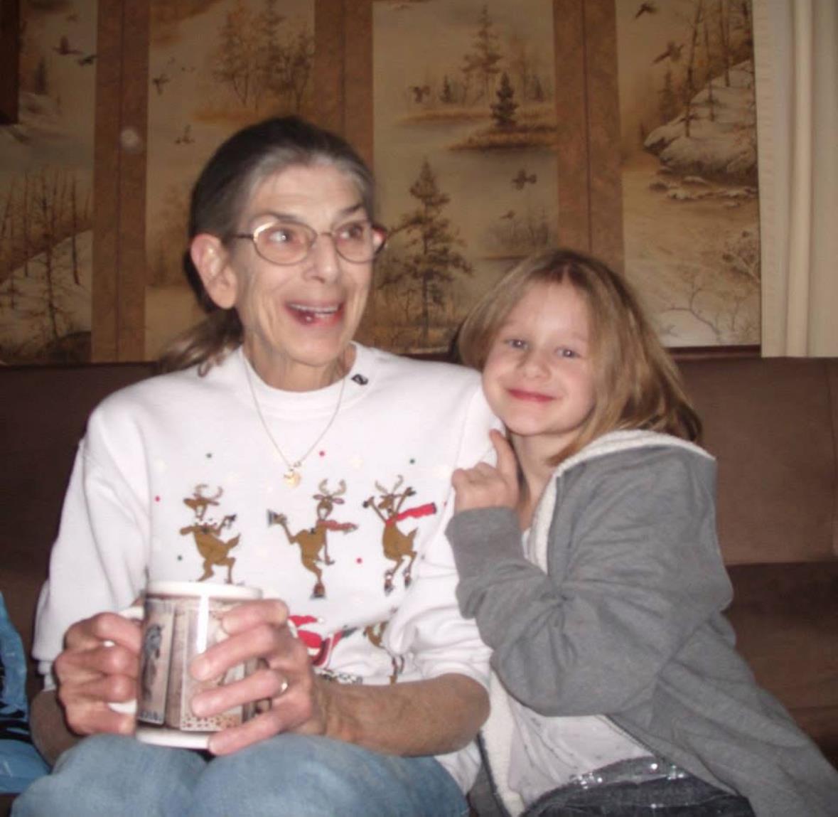 Grandmother smiles joyfully while holding a cup as her granddaughter cuddles next to her.
