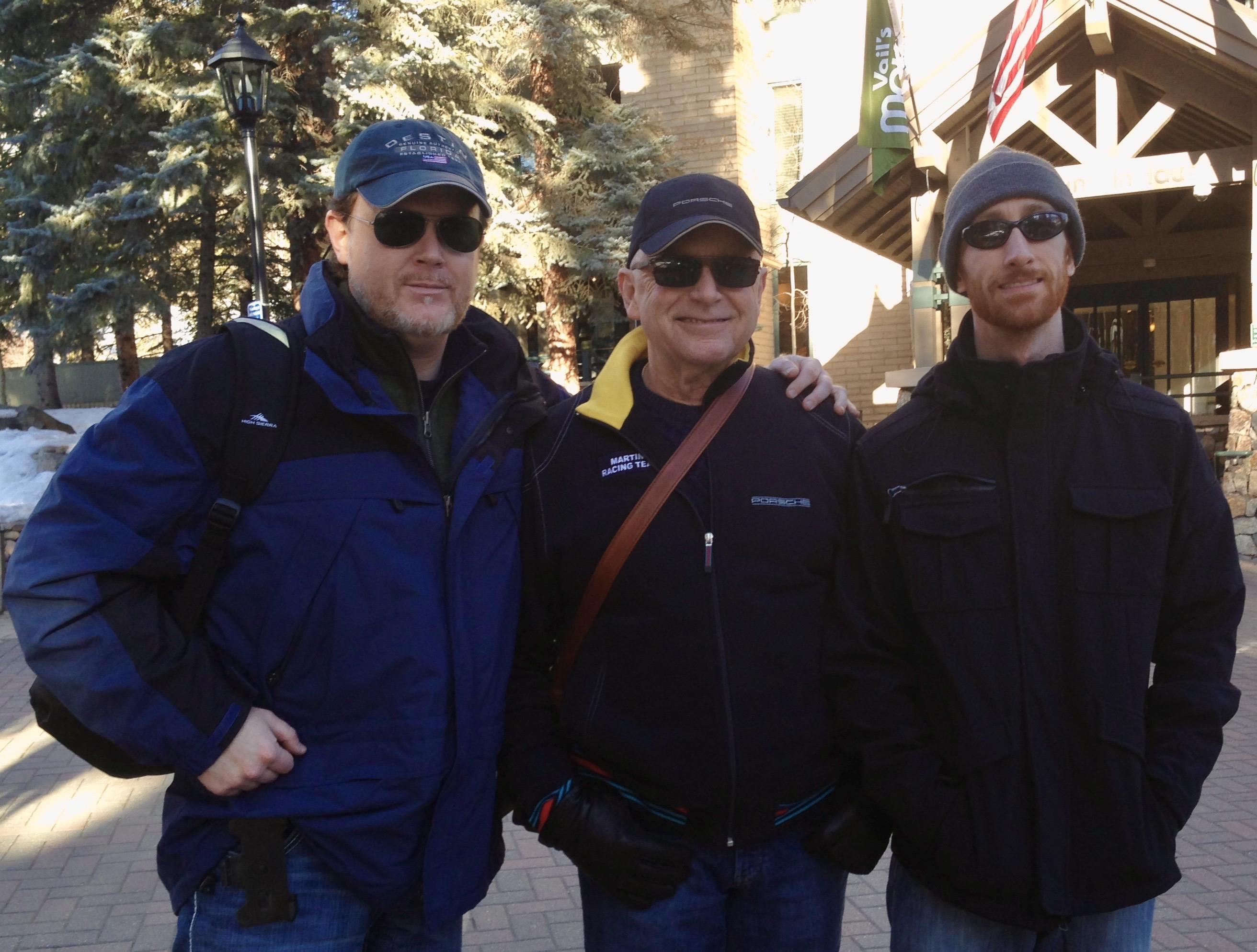Three men stand smiling outdoors, dressed in warm winter attire, surrounded by snow-covered trees.