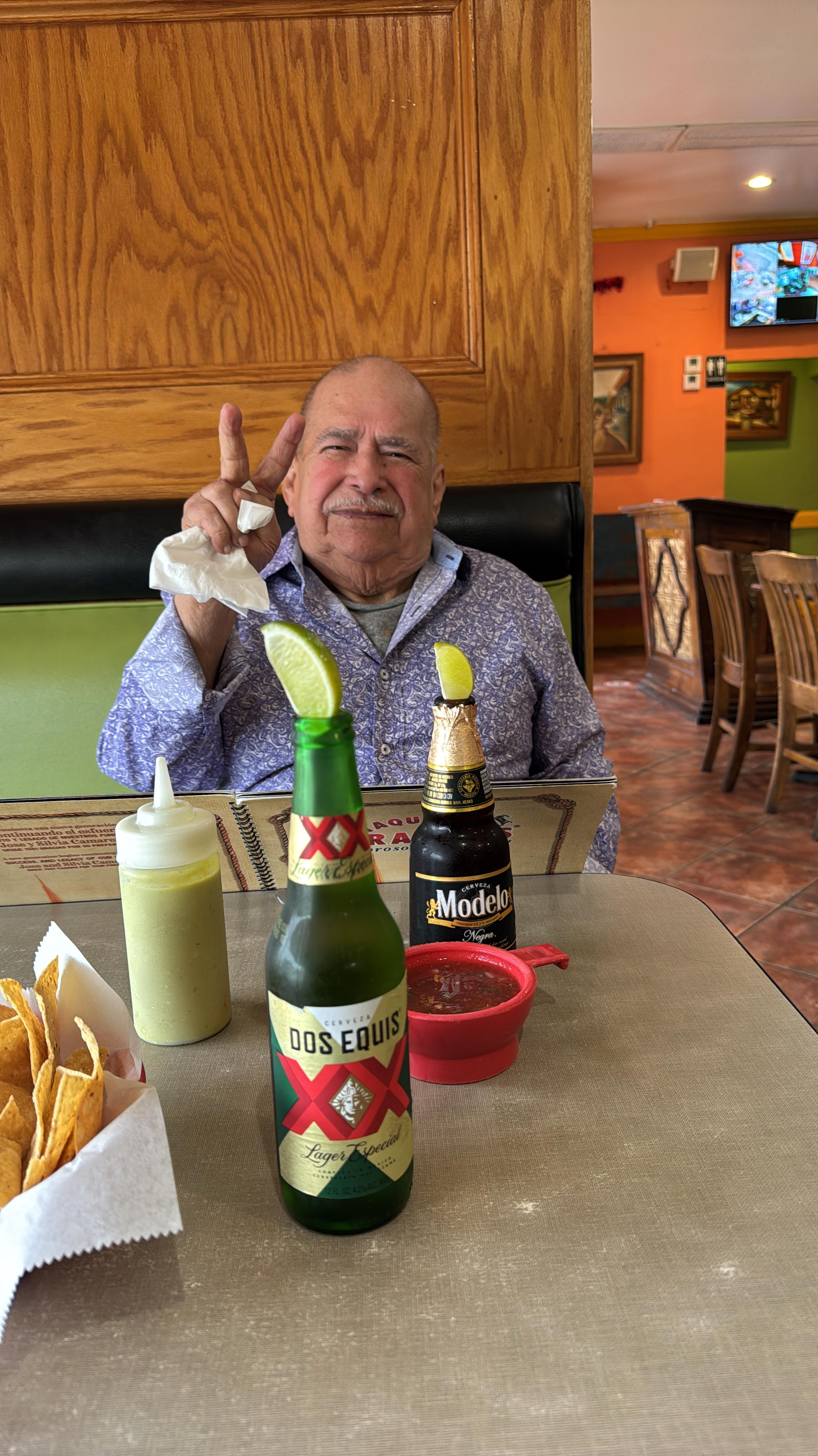 An elderly man smiles and raises two fingers while enjoying drinks at a busy restaurant.