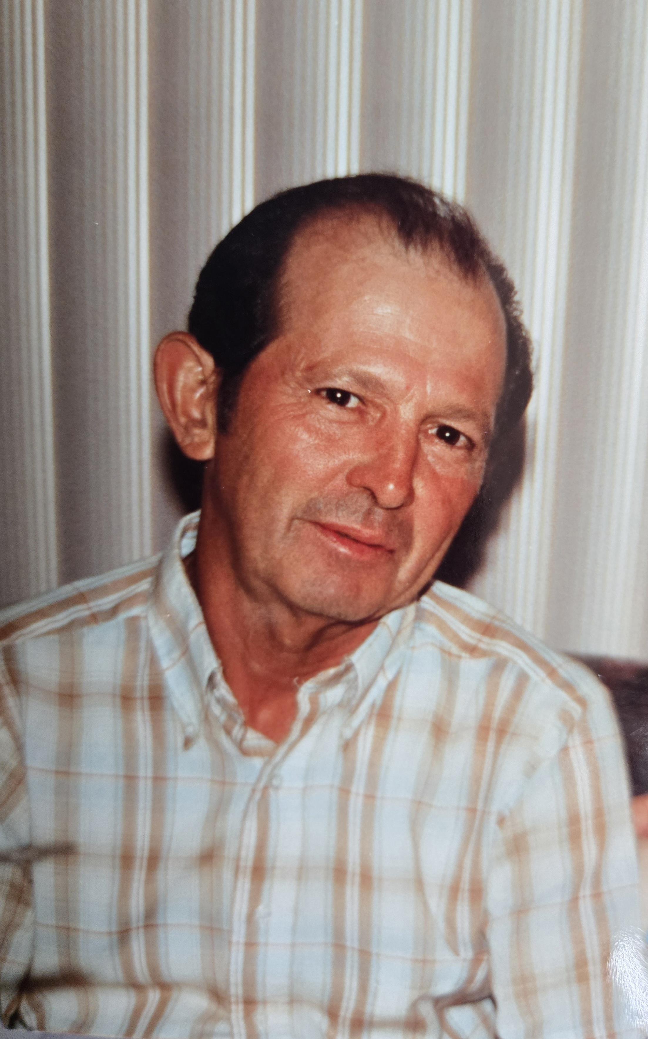 A man with a gentle smile is seated indoors, dressed in a light plaid shirt.