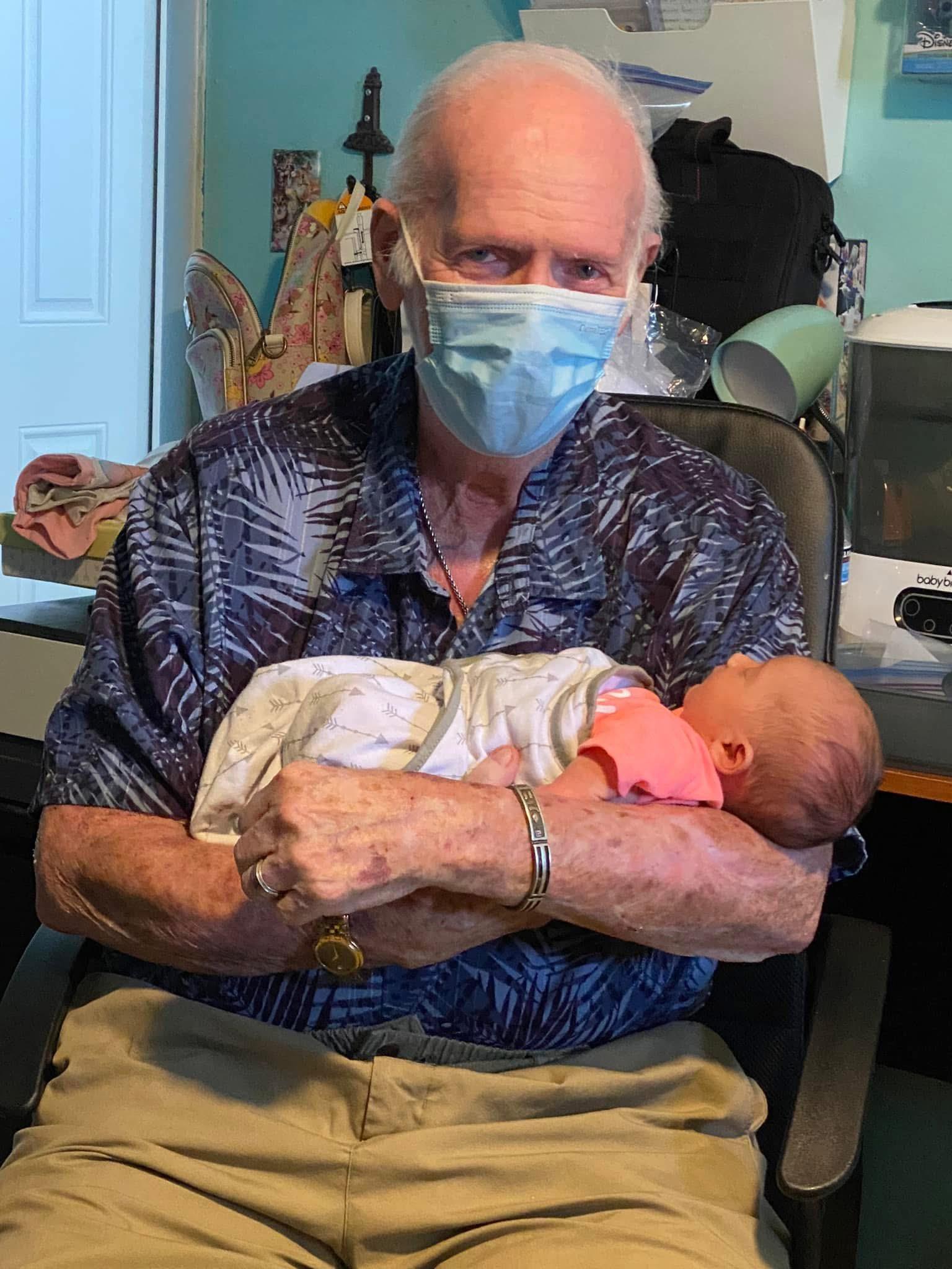 Elderly man lovingly cradles an infant in his arms while seated at home, both wearing masks.