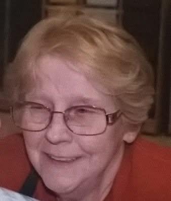 An elderly woman with short blonde hair and glasses smiles warmly while interacting with others.