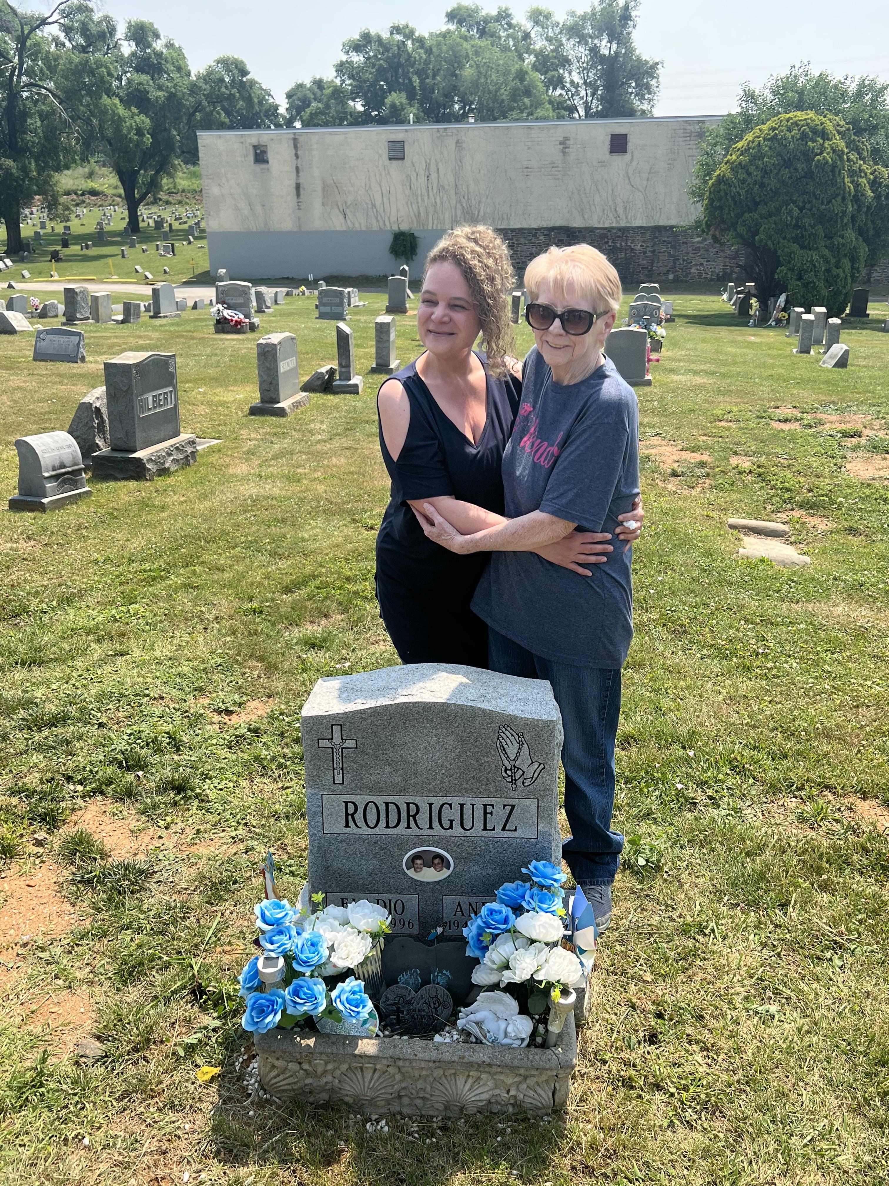 Two relatives stand together at a grave, surrounded by flowers, sharing a heartfelt moment.