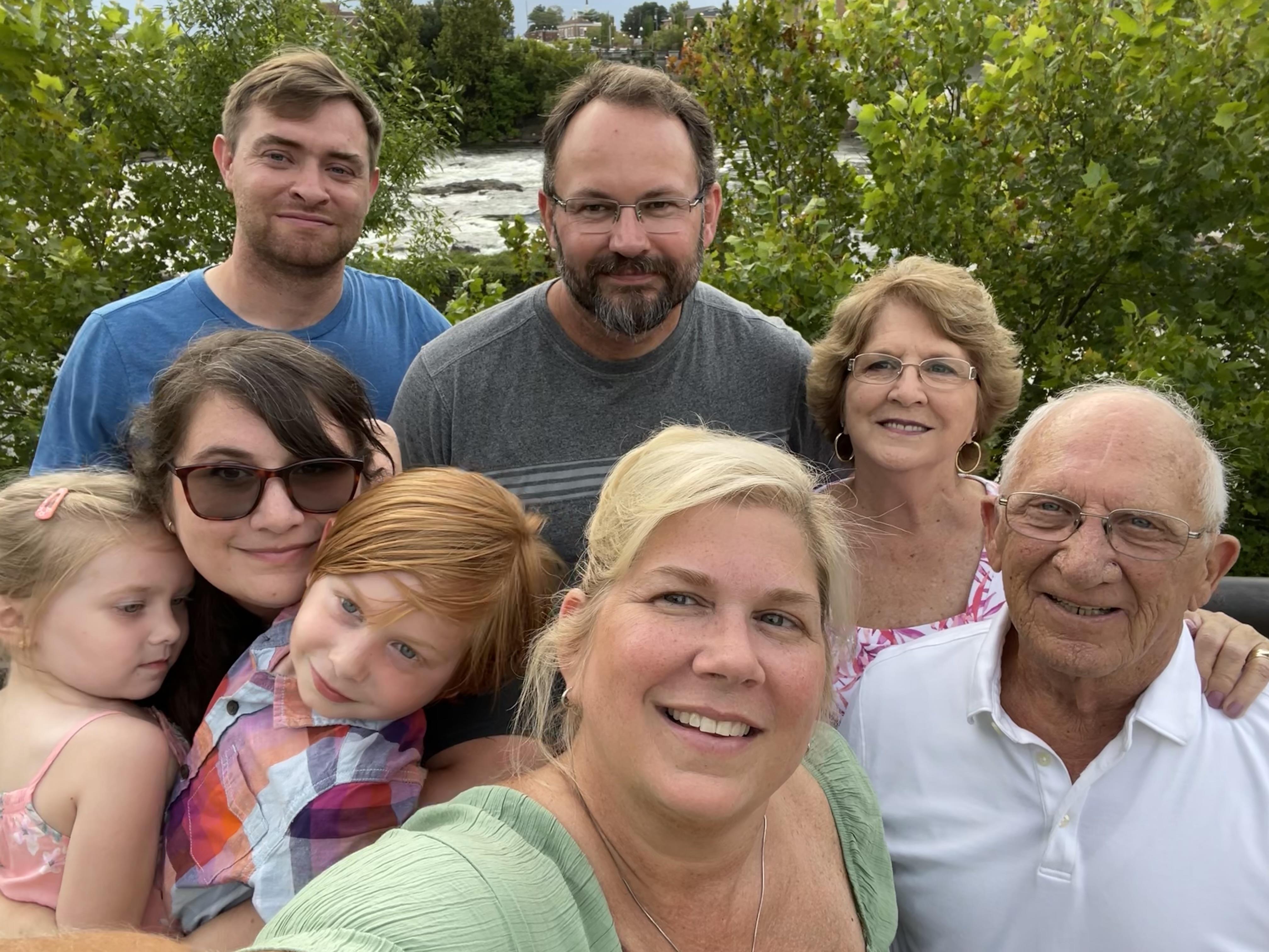 A joyful family poses together in front of a river, enjoying a sunny day outdoors with smiles.