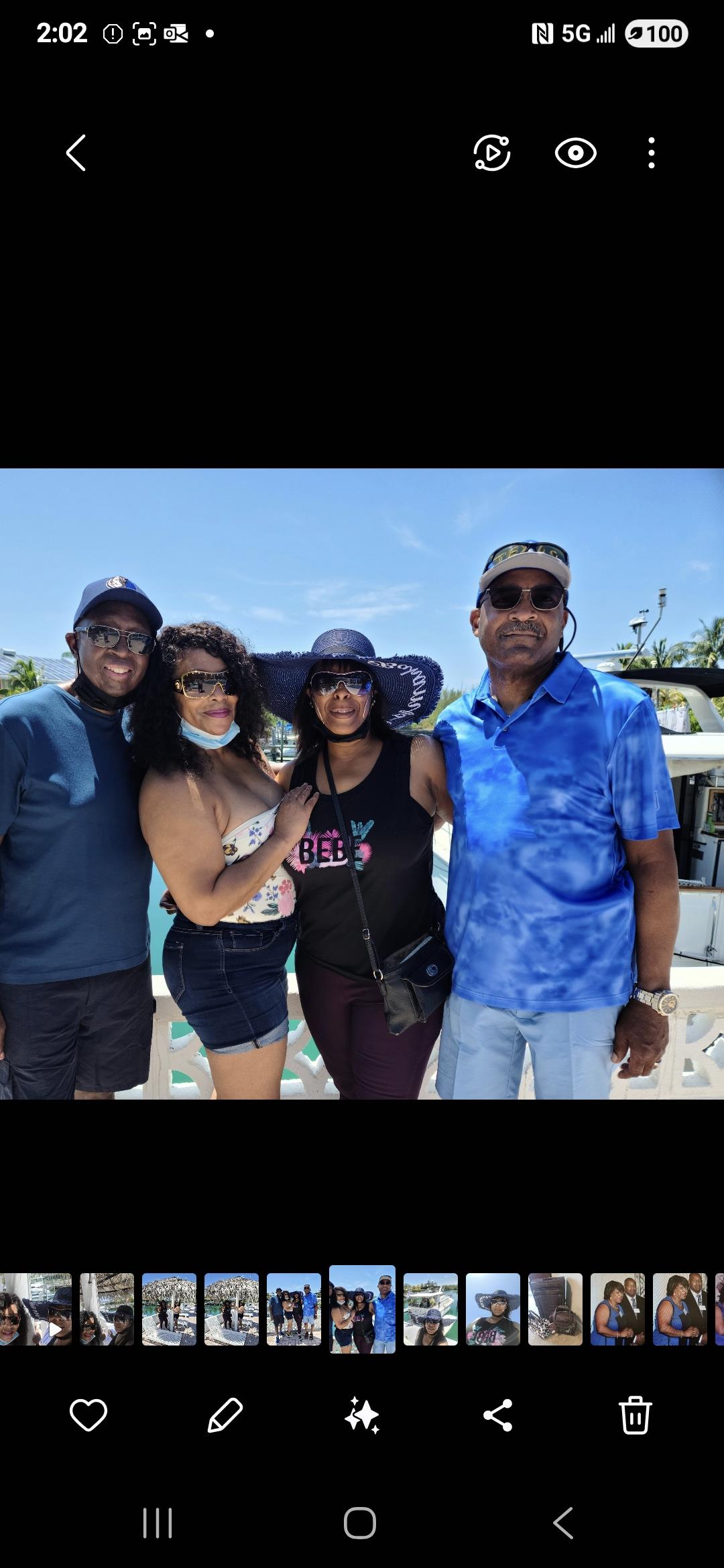 Four friends smile together in bright sunlight at the beach, dressed in summer attire.