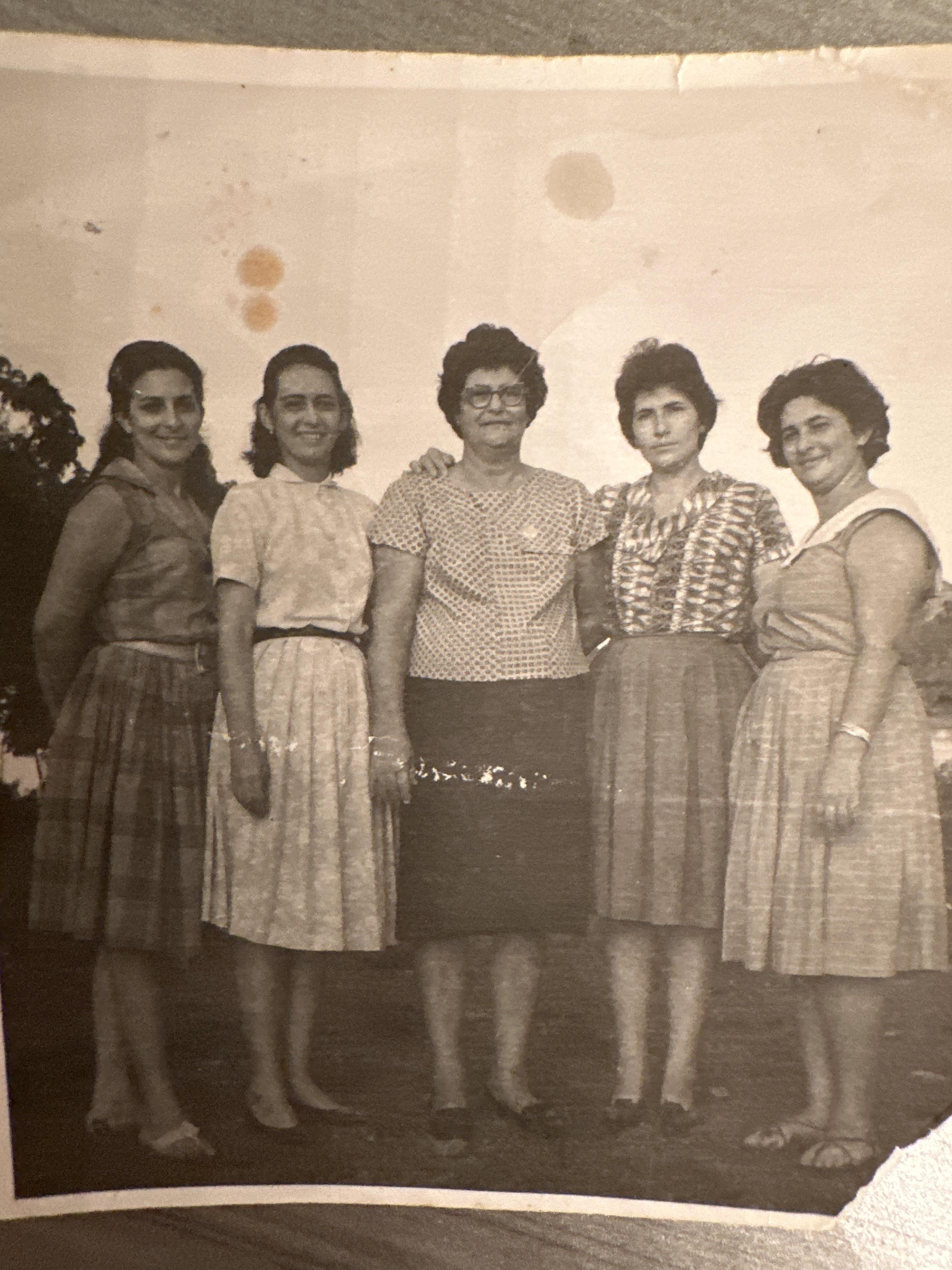Five women pose outdoors with smiles, dressed in vintage dresses, enjoying a sunny day.