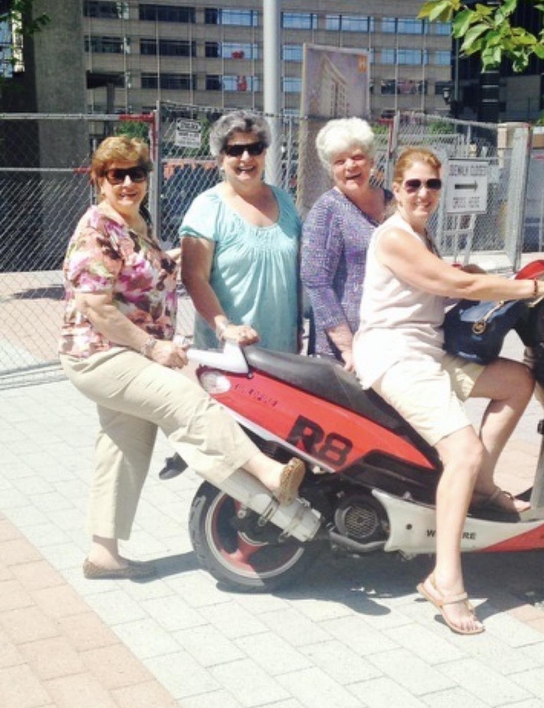 Four women smile and pose on a scooter in a lively urban environment under bright sunlight.