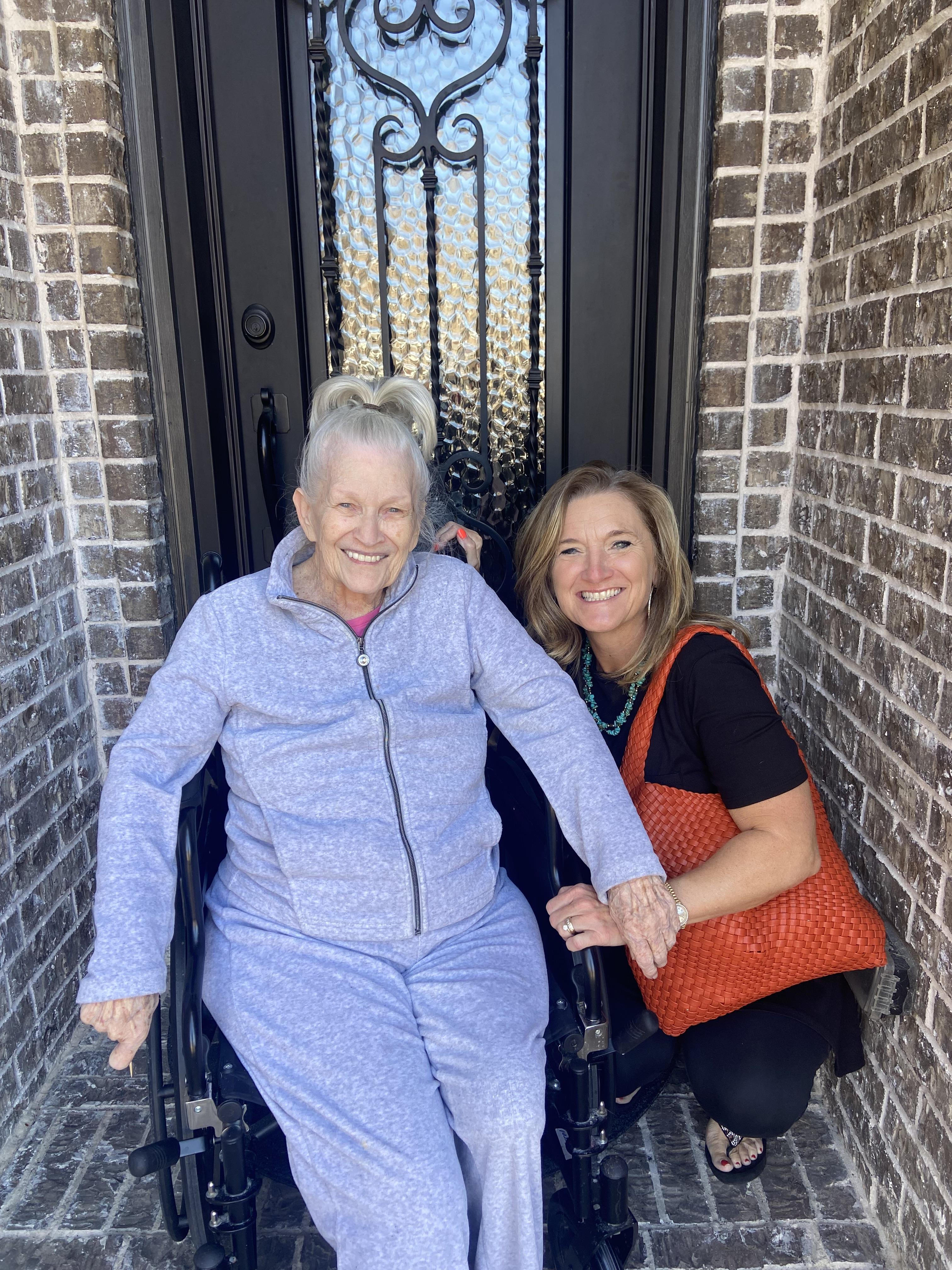 Two women enjoy a warm moment at a front door, one in a wheelchair, smiling brightly together.