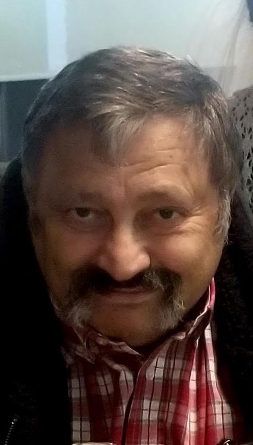 Man with a friendly smile poses indoors, showcasing his mustache and plaid shirt in a cozy setting.