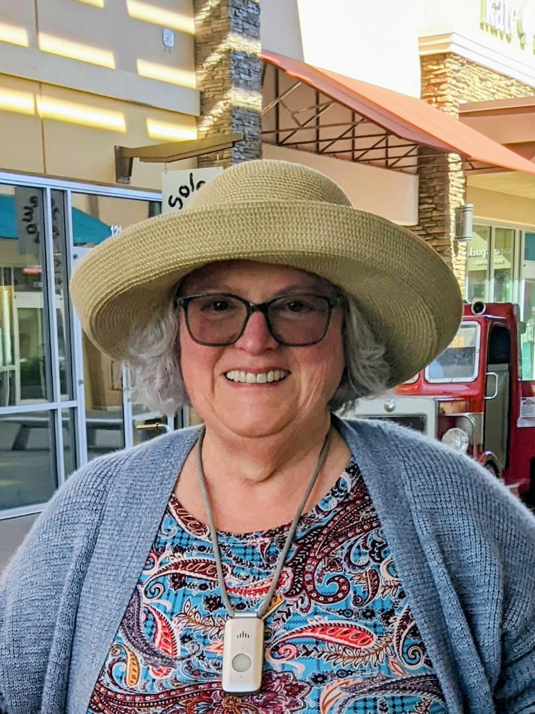 A cheerful woman wearing glasses and a hat poses at an outdoor shopping area during the day.