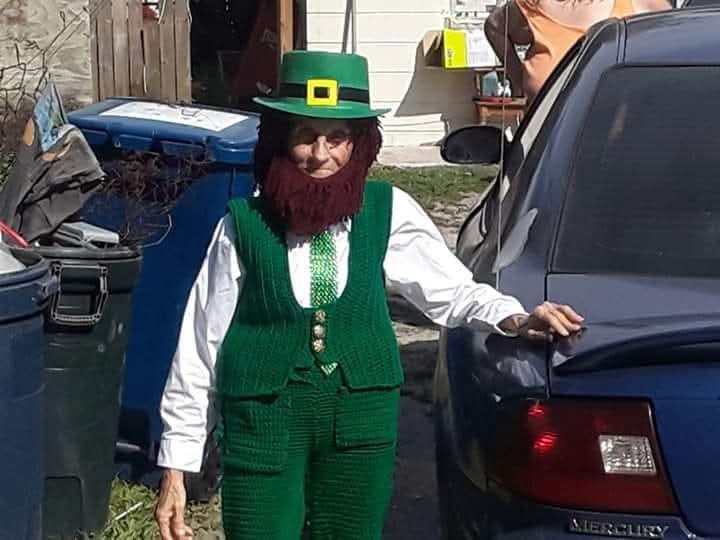 An elderly individual in a green leprechaun costume poses outdoors beside a parked car.