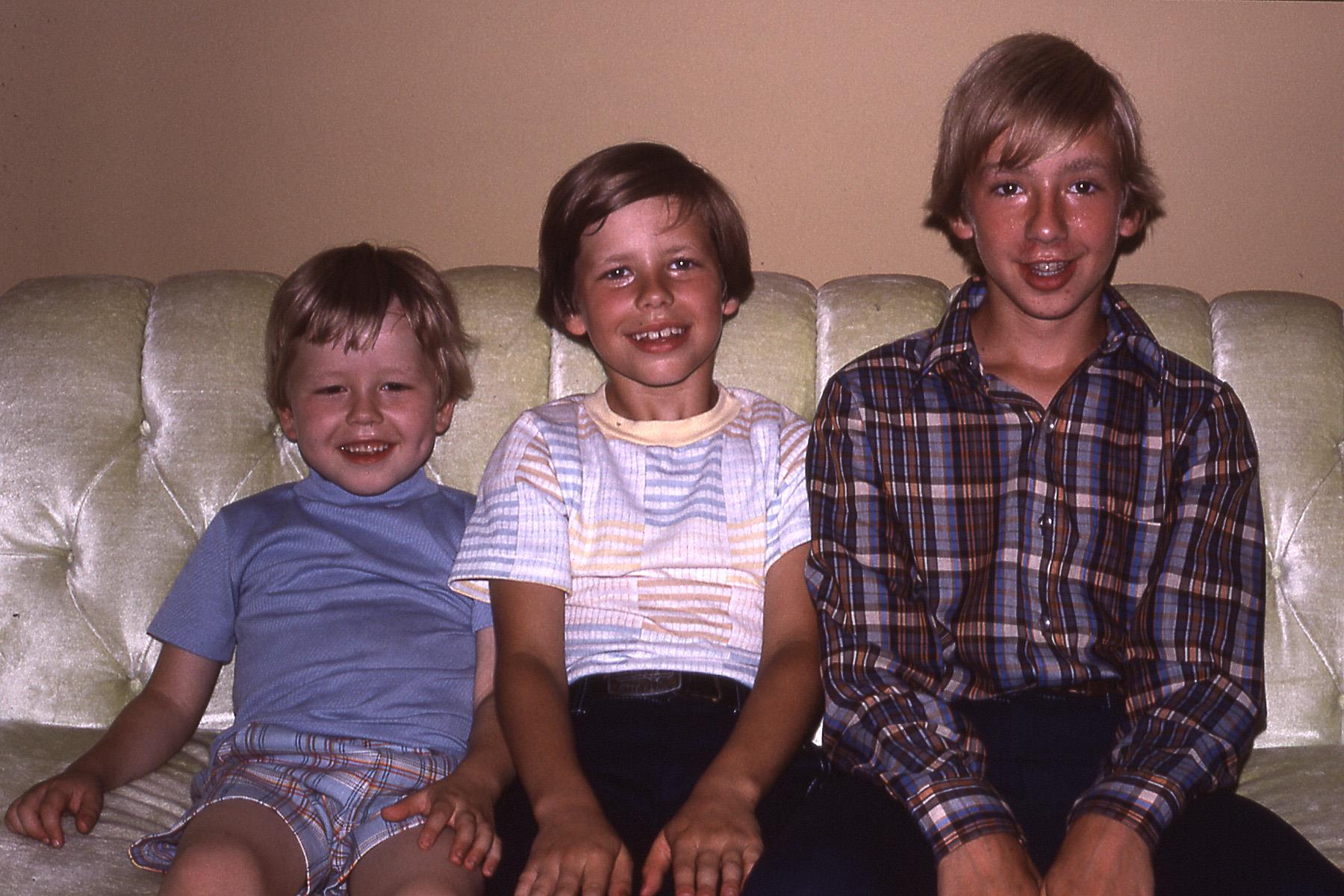 Three boys sit closely, smiling happily on a green couch in a cozy living room setting.