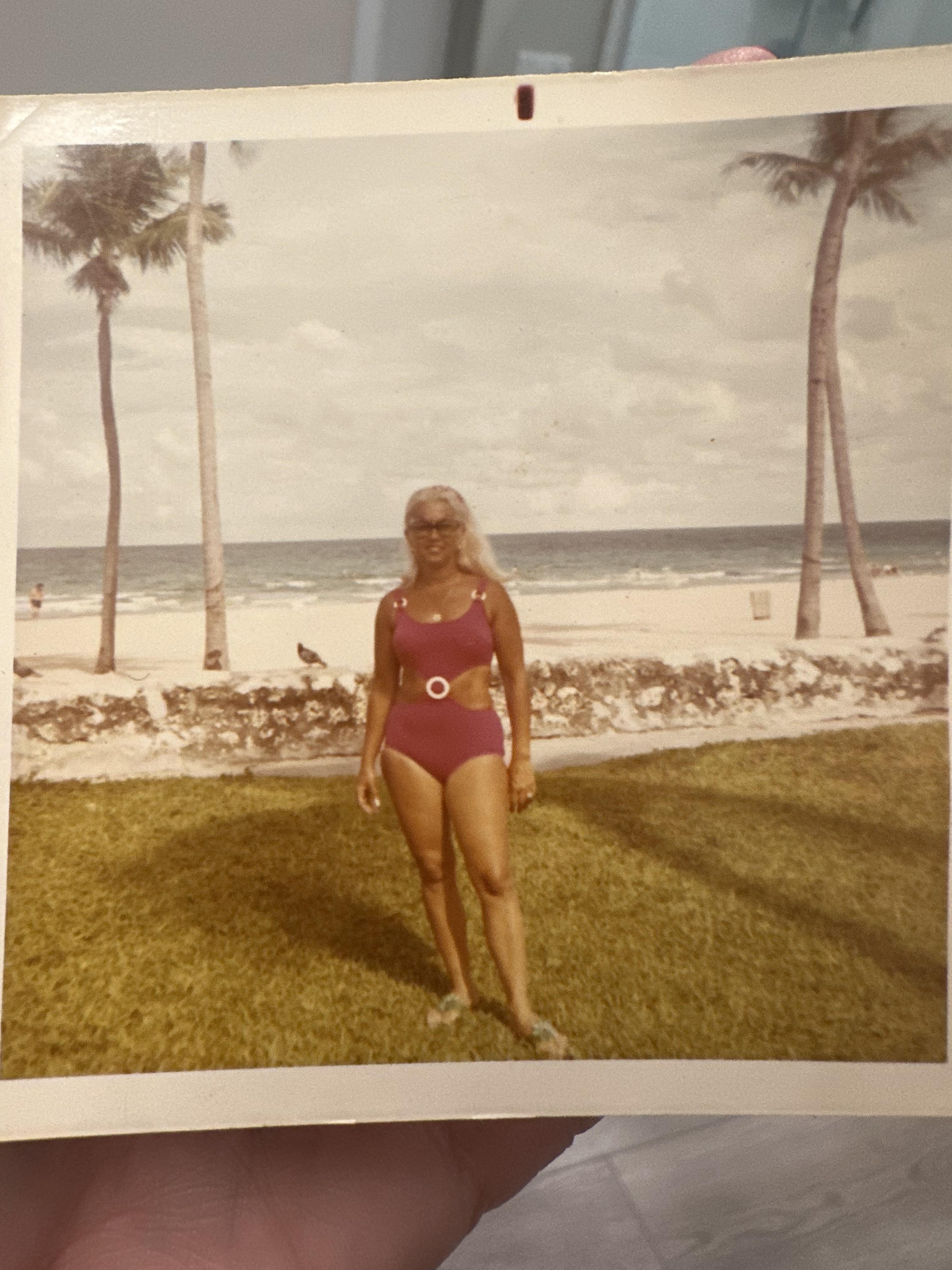 A woman poses in a pink swimsuit on grassy beach area under palm trees by the ocean.