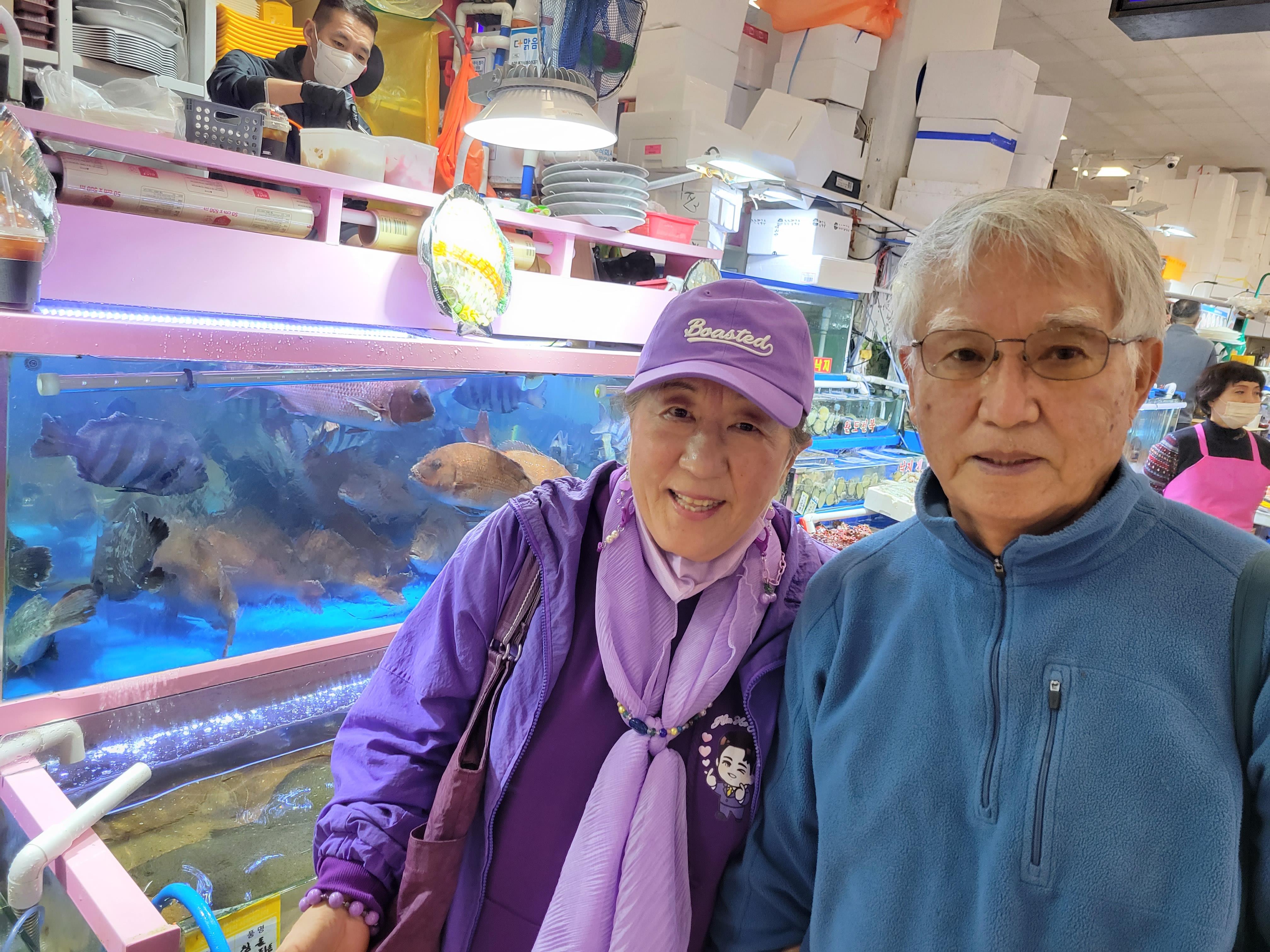 An elderly couple smiles for the camera at a lively fish market with colorful tanks.