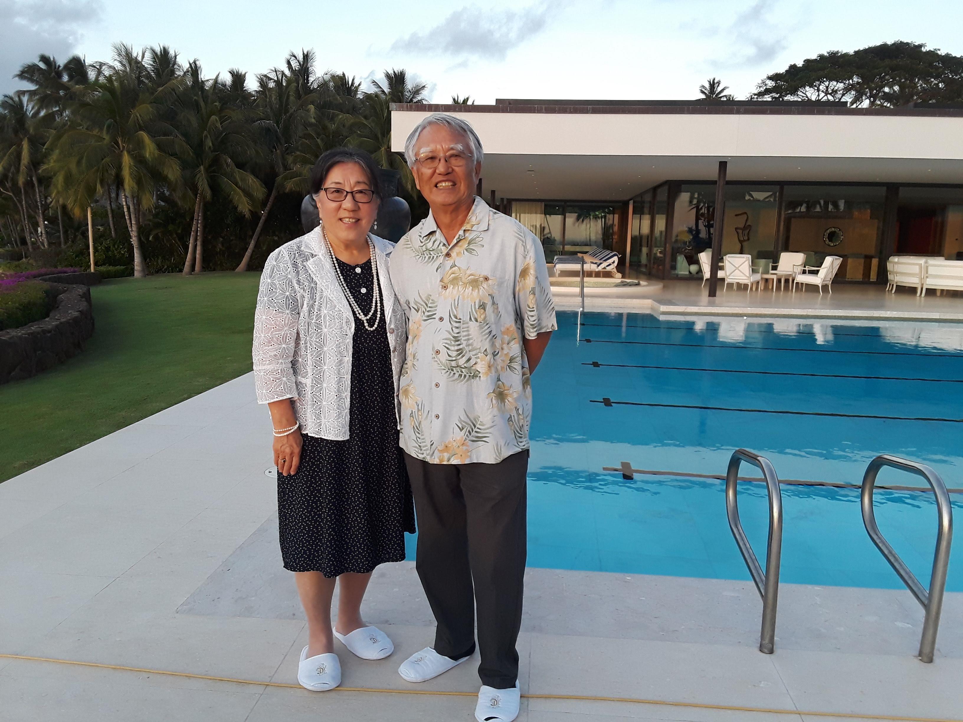 An elderly couple stands together at a poolside, surrounded by palm trees during sunset.