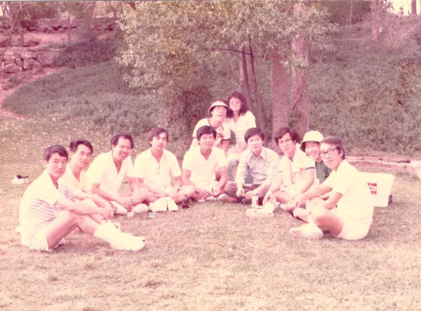 Friends gather on the grass for a relaxing picnic in a park surrounded by trees.