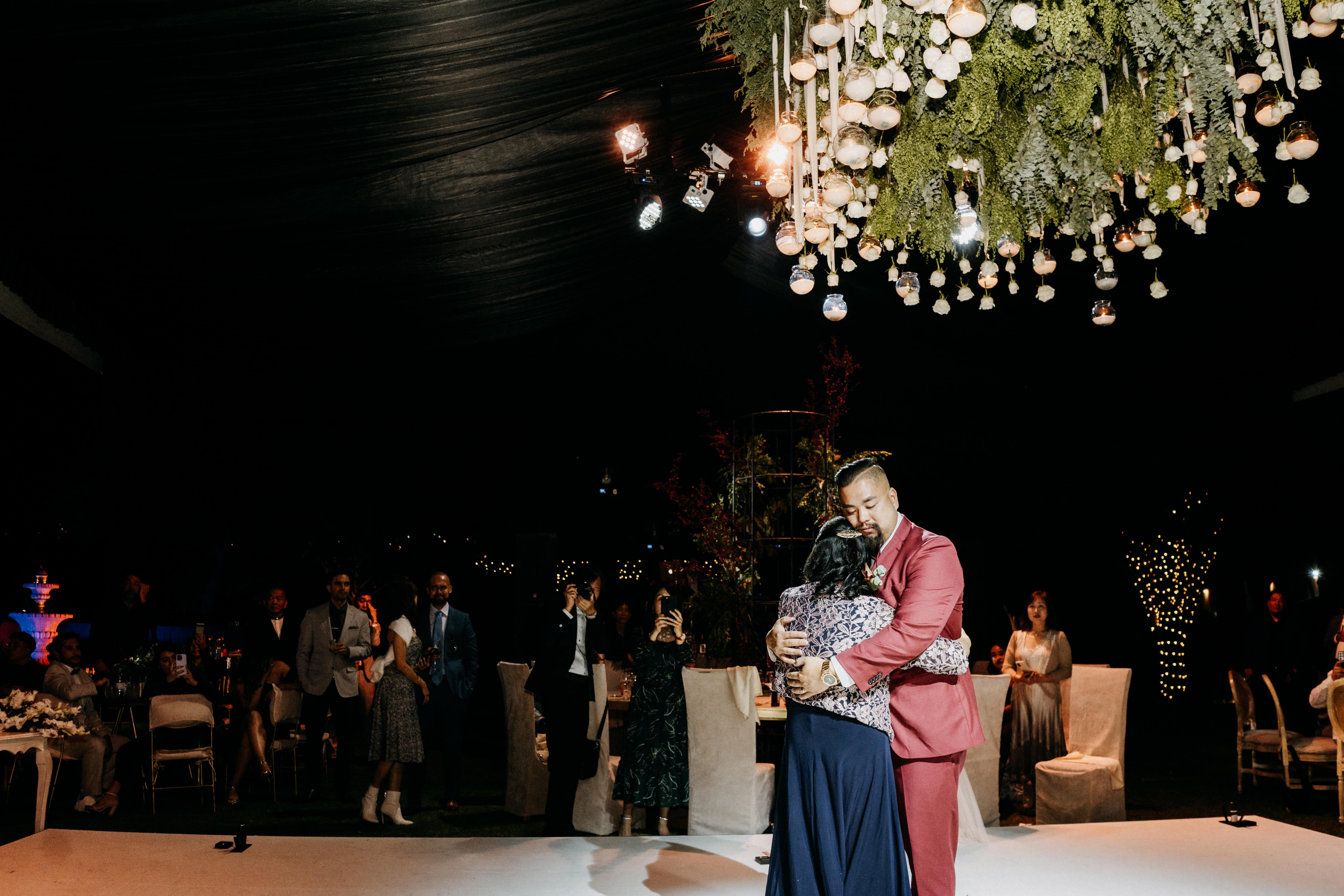 A couple embraces while dancing under hanging lights at a stylish evening event with guests nearby.