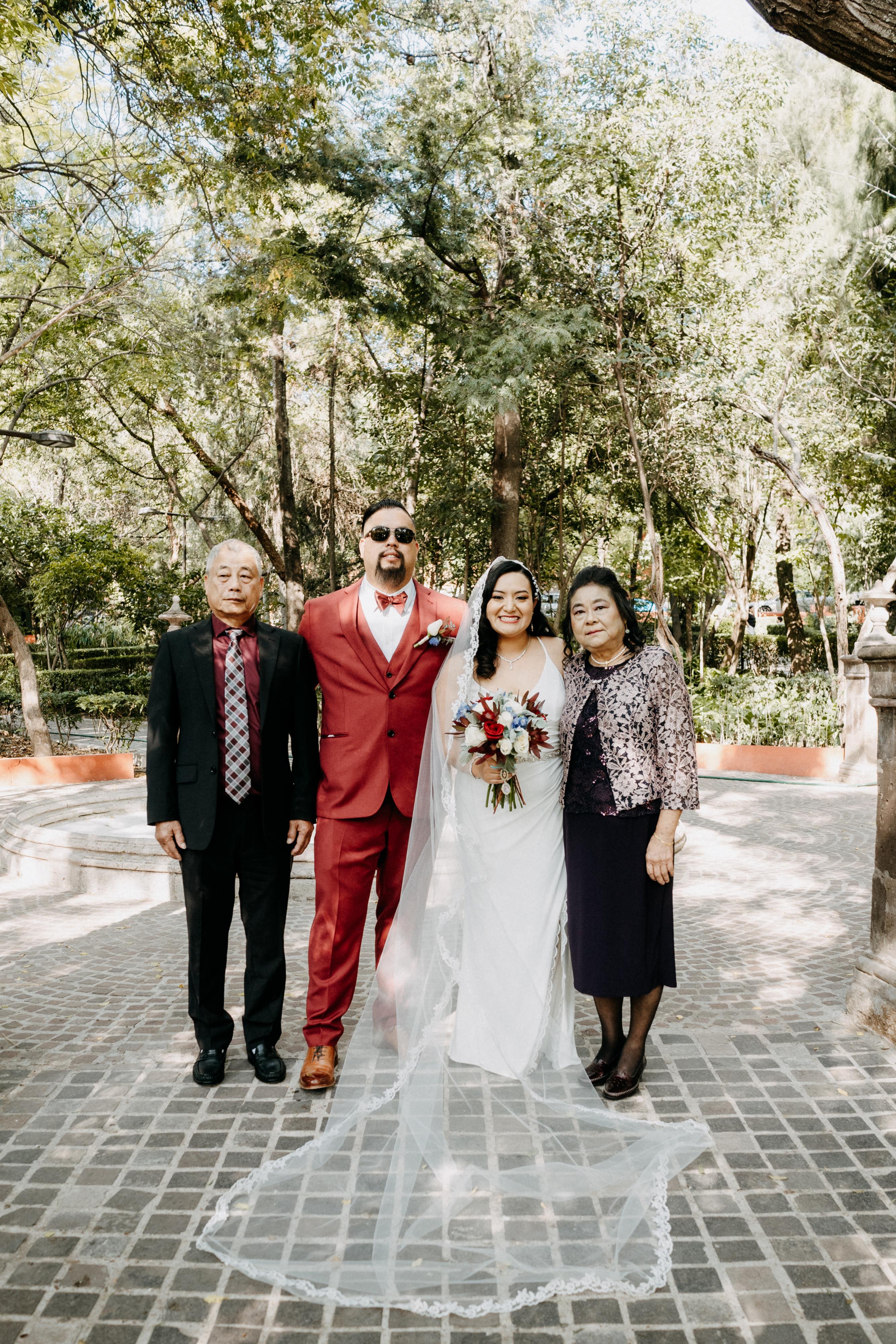 Newlywed couple poses happily with their parents in a beautiful lush park setting.