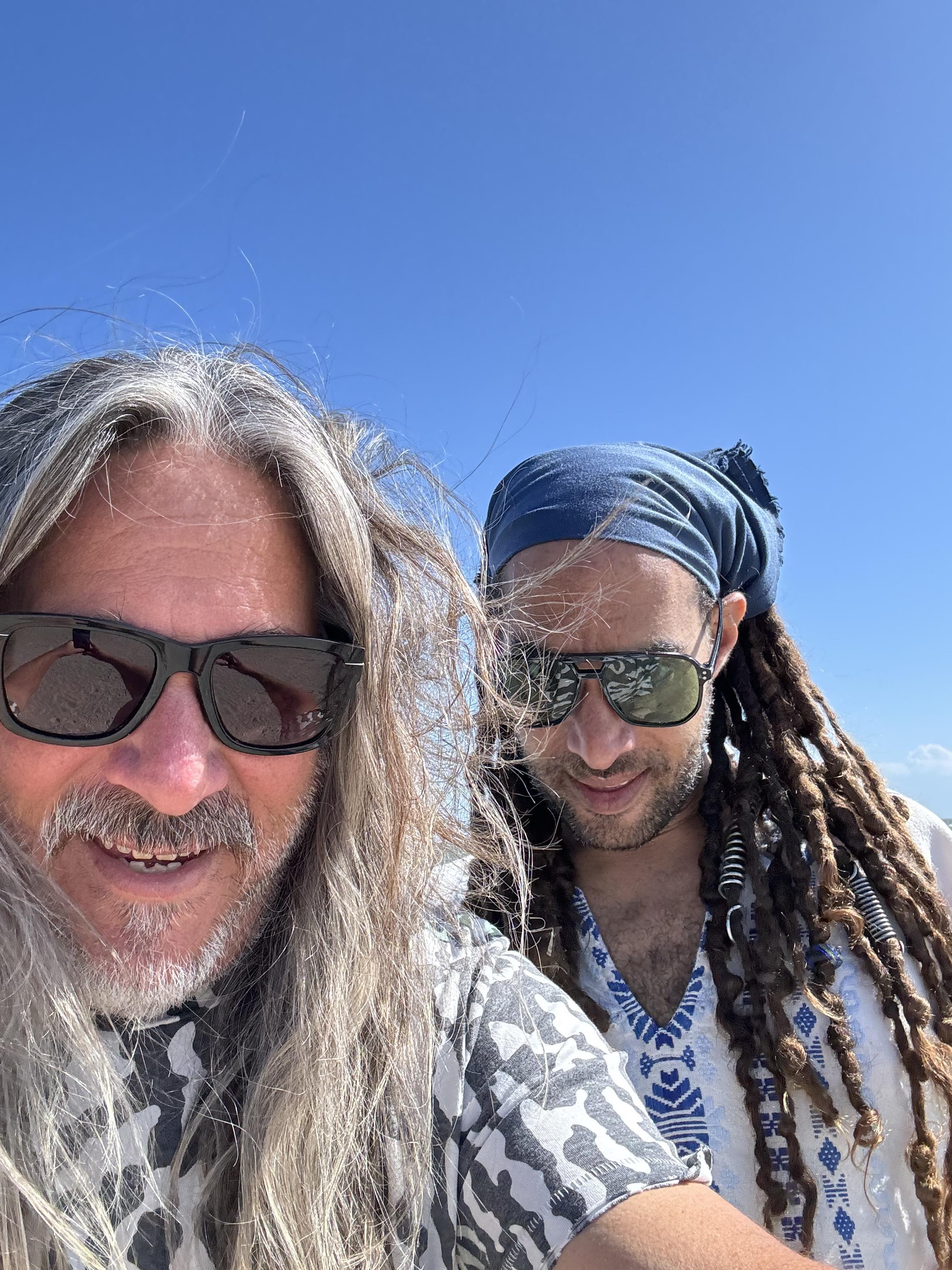 Two friends pose together on a beach, enjoying the sun, with longer hair flowing in the breeze.