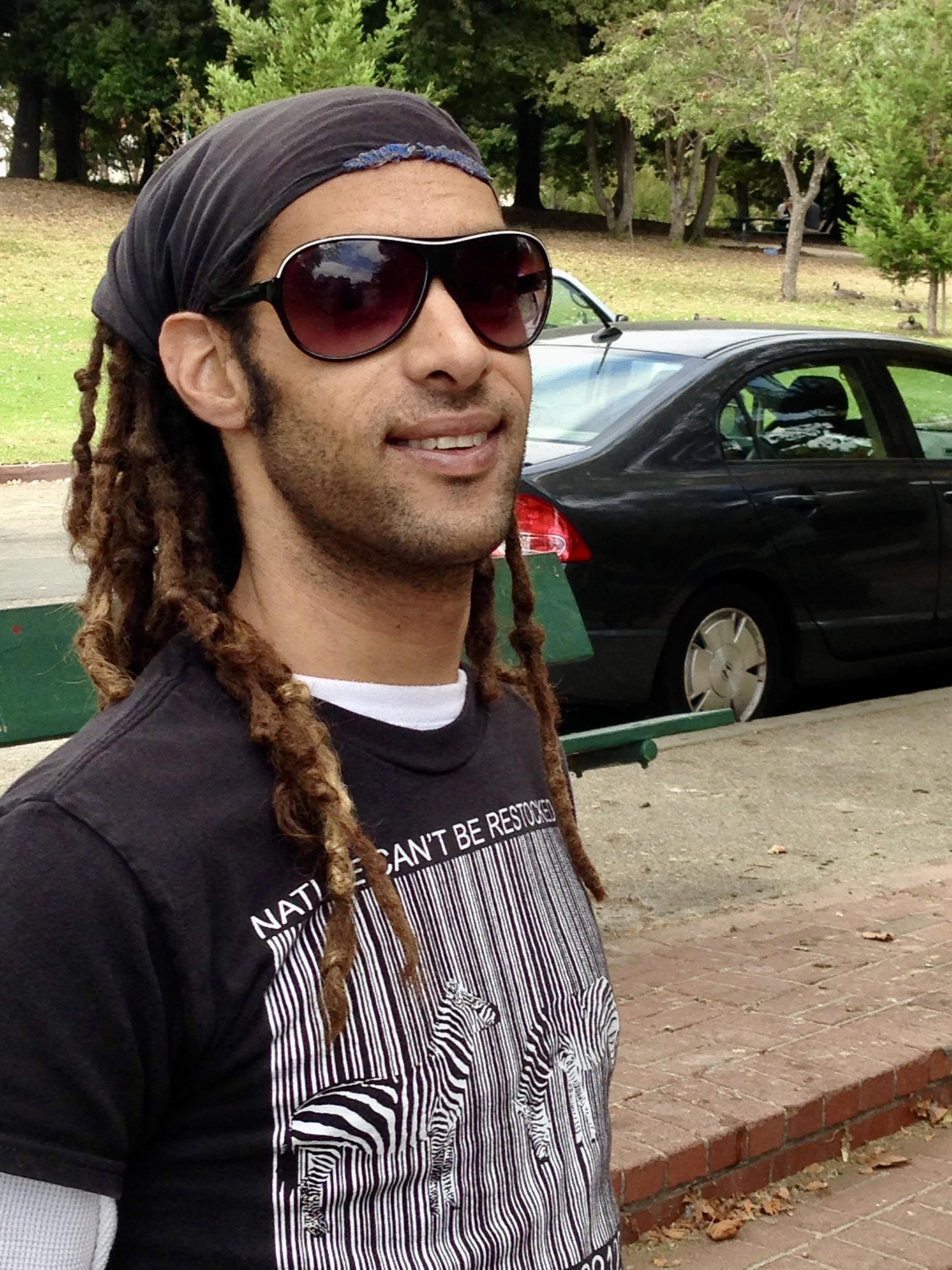 A man with long dreadlocks and sunglasses stands in a park, smiling and enjoying the sunlight.