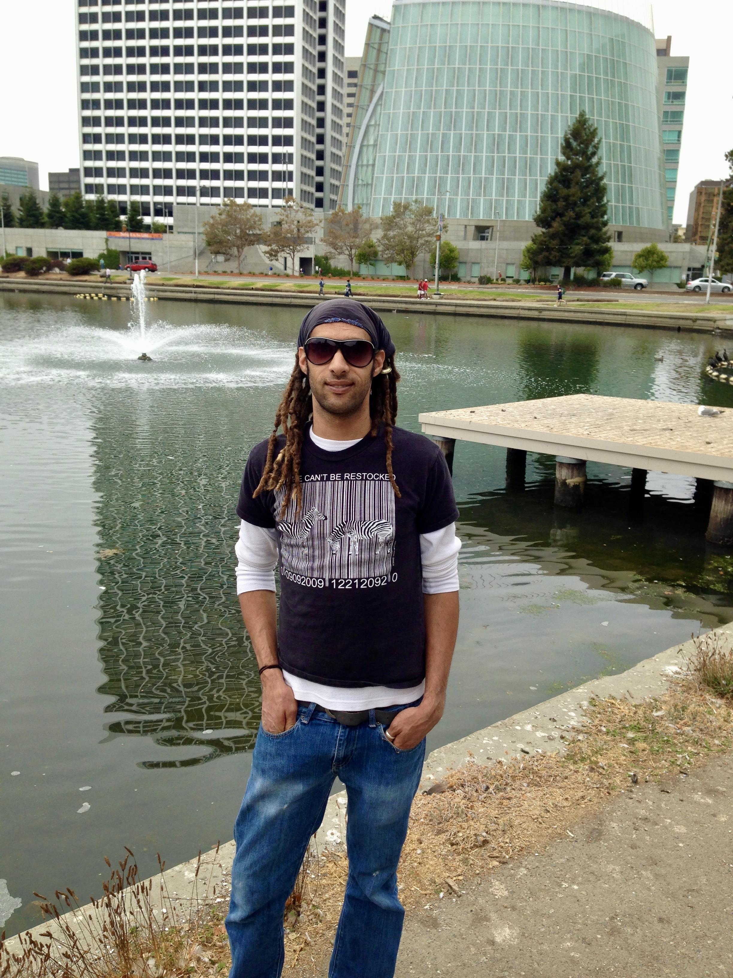 A man with long hair poses confidently near a lake surrounded by modern architecture.