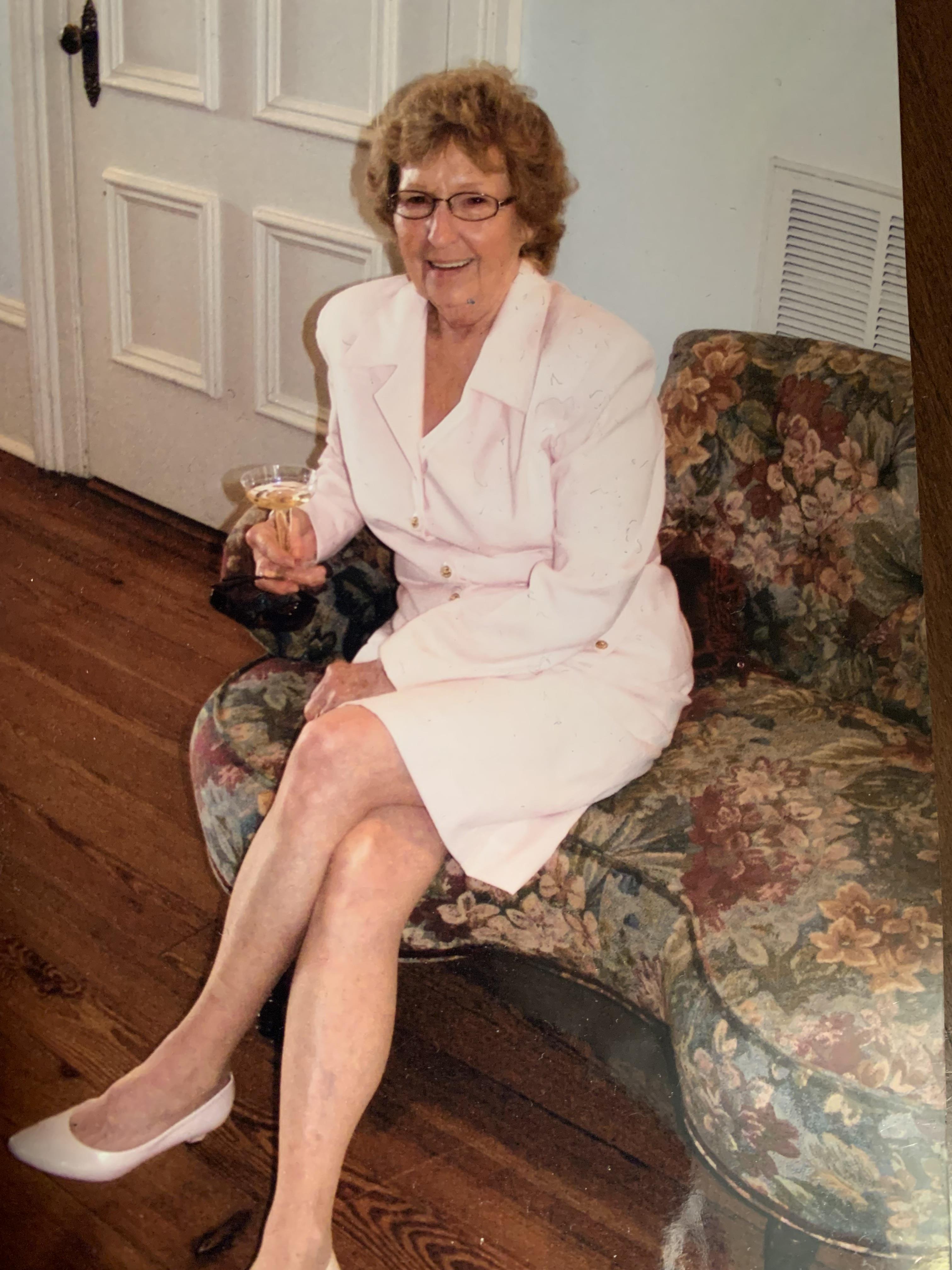 An elderly woman in pink sits on a floral couch with a drink in a cozy living room.