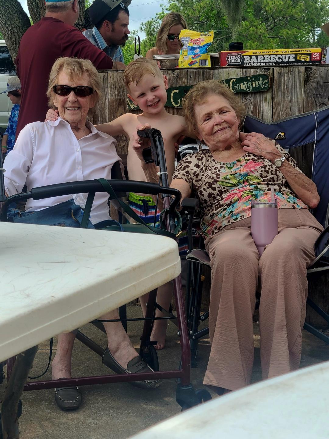 A smiling boy poses between two elderly women in chairs at a relaxed outdoor gathering.