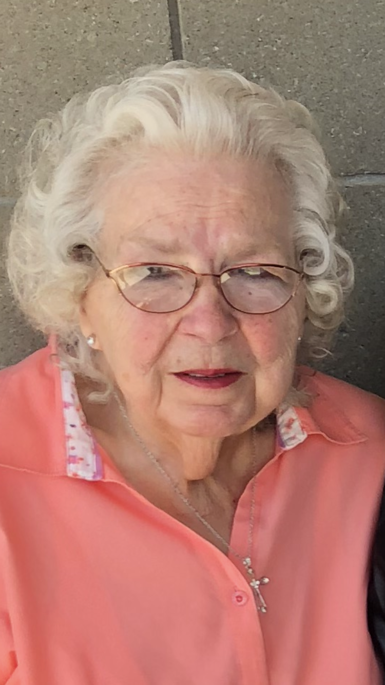 An elderly woman with white curly hair and glasses poses for a picture, exuding warmth and kindness.