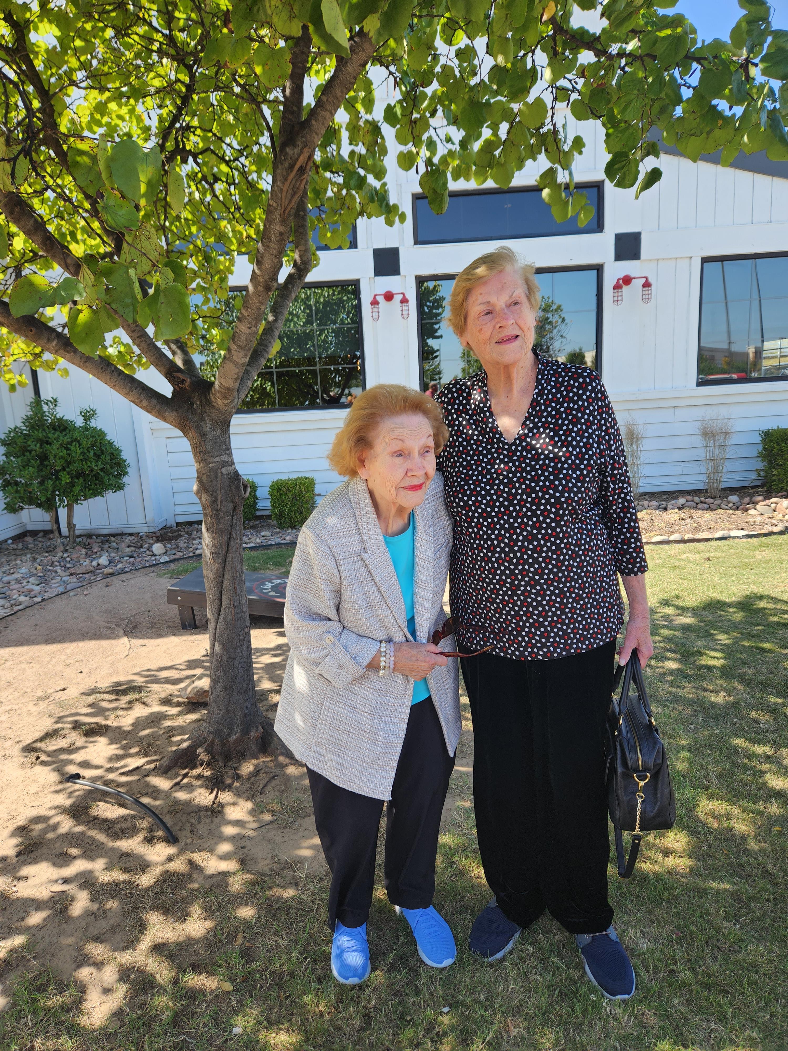 Two senior women stand together under a tree, smiling and enjoying a sunny day outdoors.