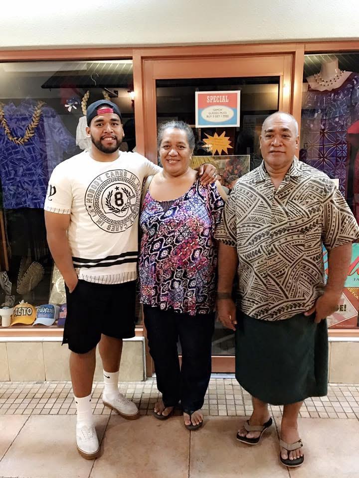 Three people pose together outside a shop, smiling warmly at the camera in a tropical area.