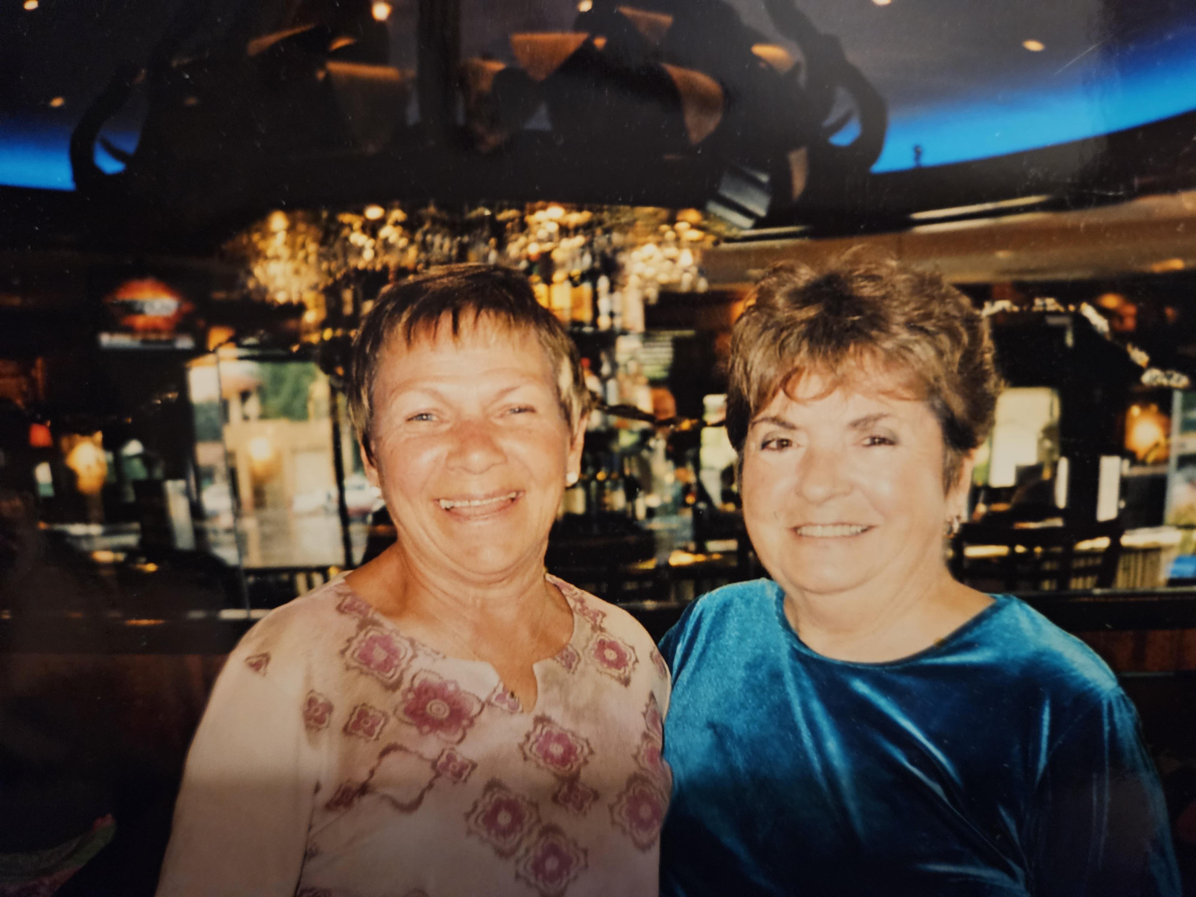 Two friends stand close, smiling happily in a lively restaurant decorated with elegant lighting.