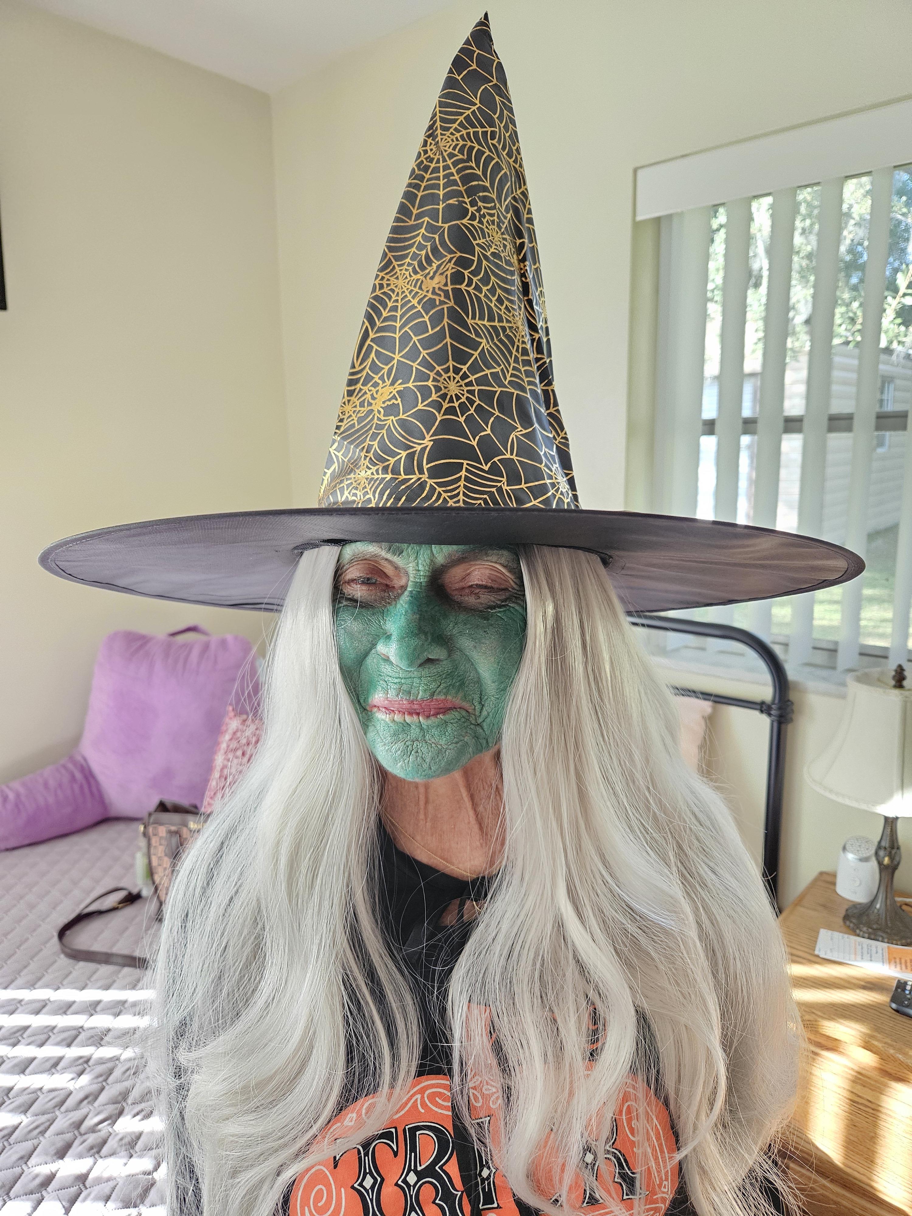 Elderly woman with long gray hair dressed as a witch enjoys Halloween festivities indoors.