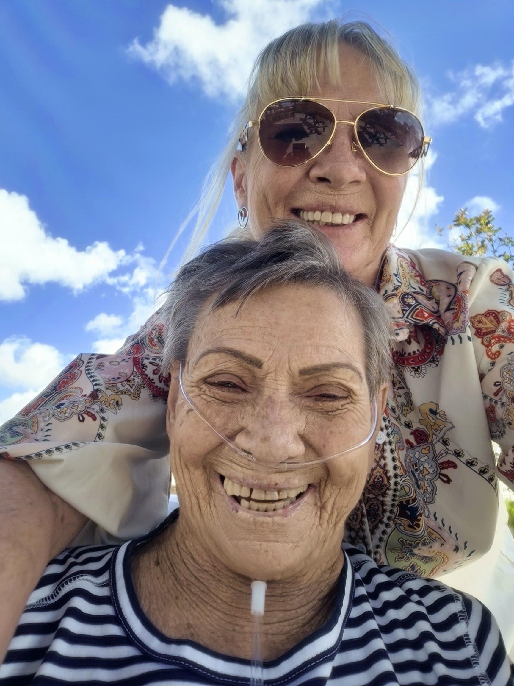 Two women are smiling broadly while enjoying a sunny day outdoors, showing joy and companionship.