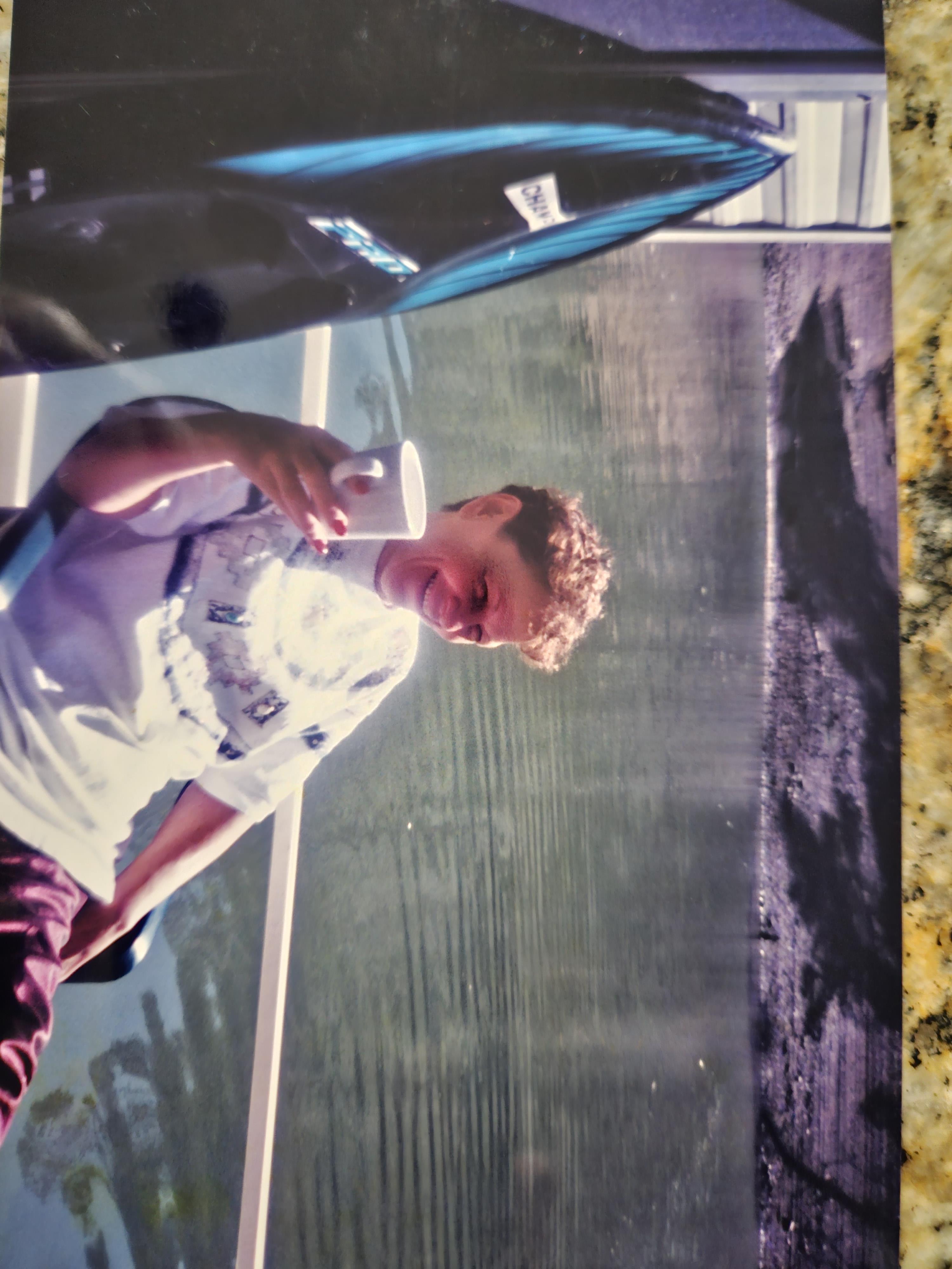Boy holds a cup and smiles while resting on a boat beside calm water on a bright day.