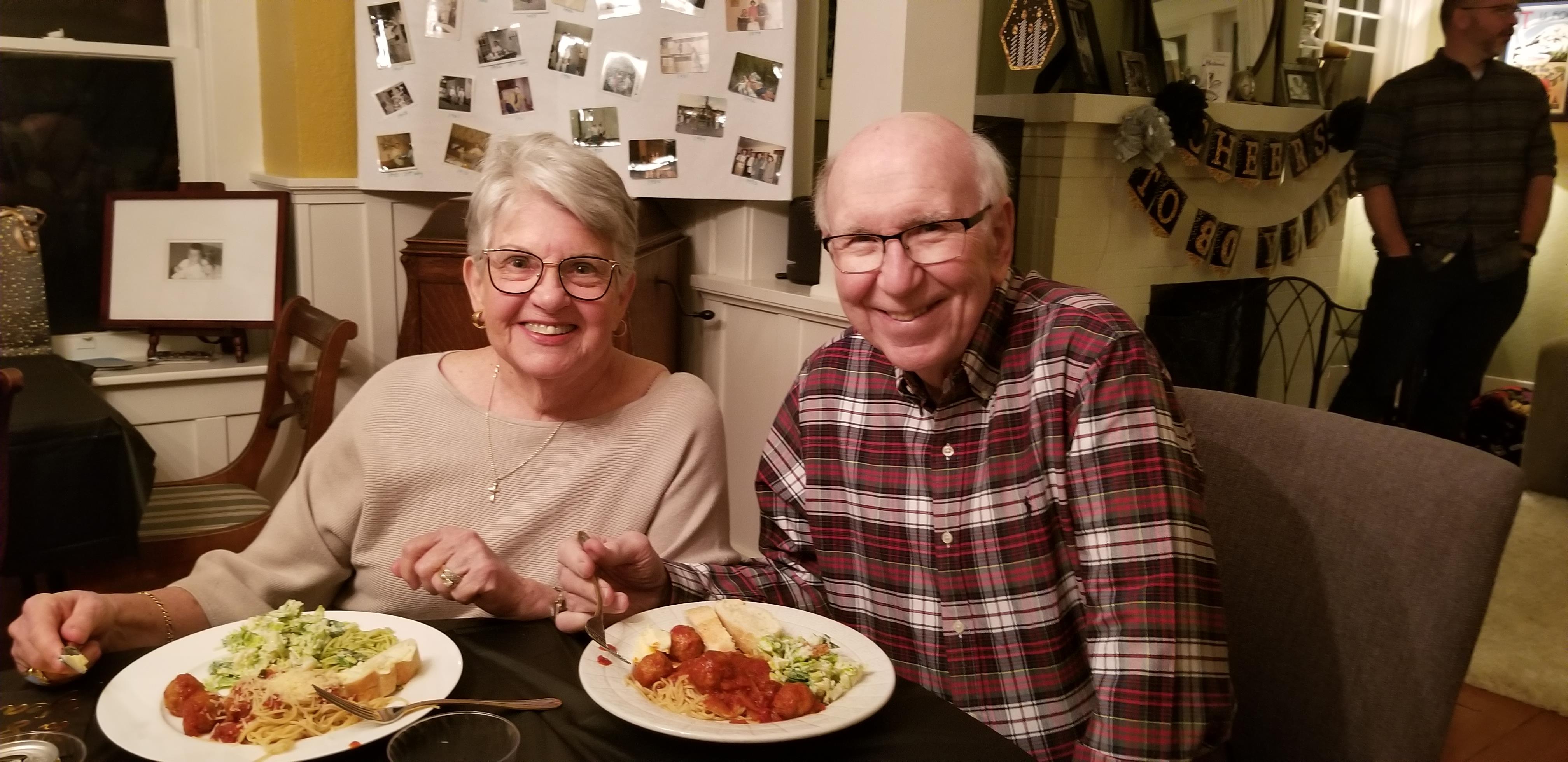 An elderly couple sits at a dining table, happily sharing a meal together in their home.