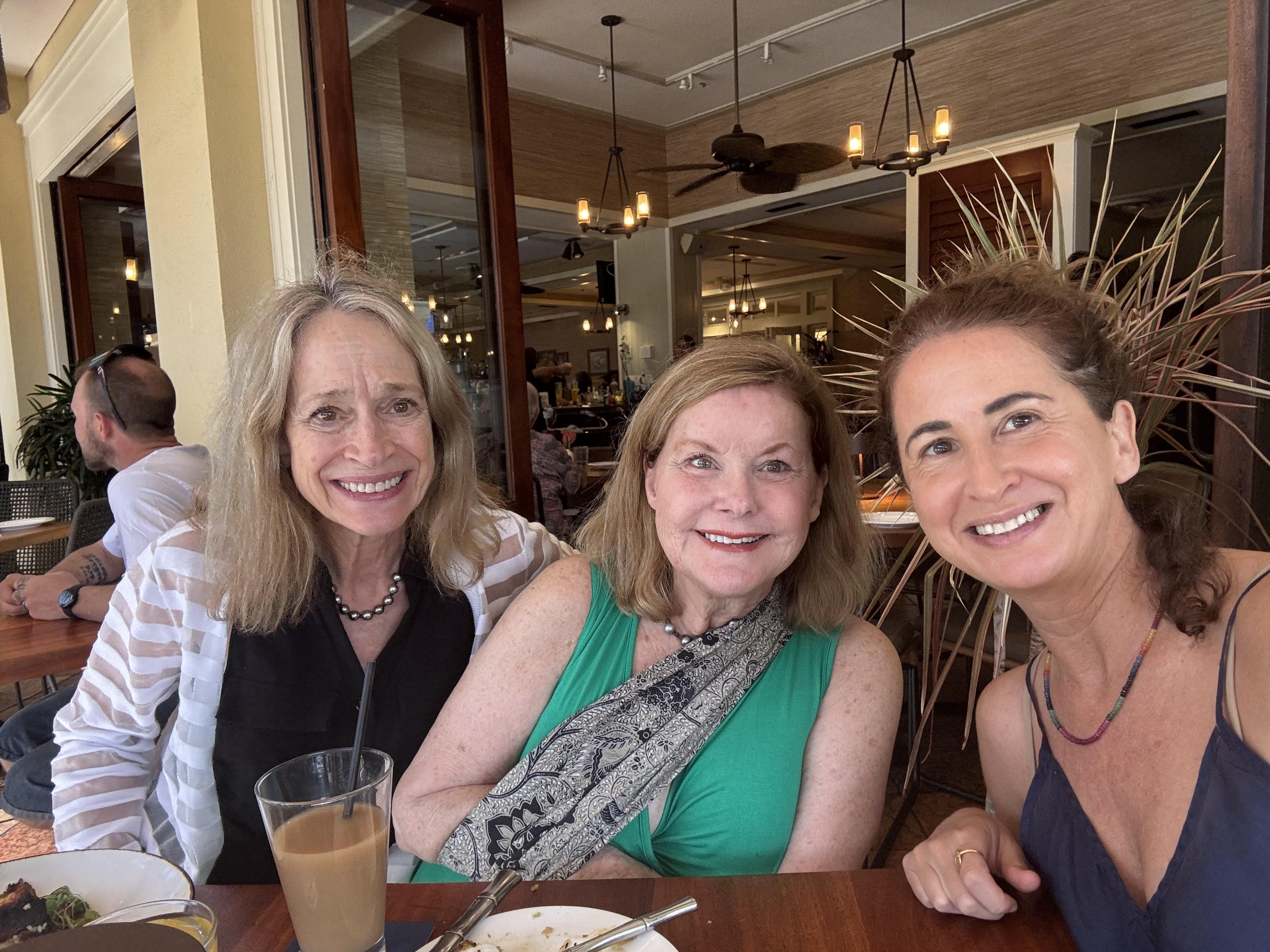 Three women gather around a table, sharing smiles and drinks while enjoying lunch in a lively café.