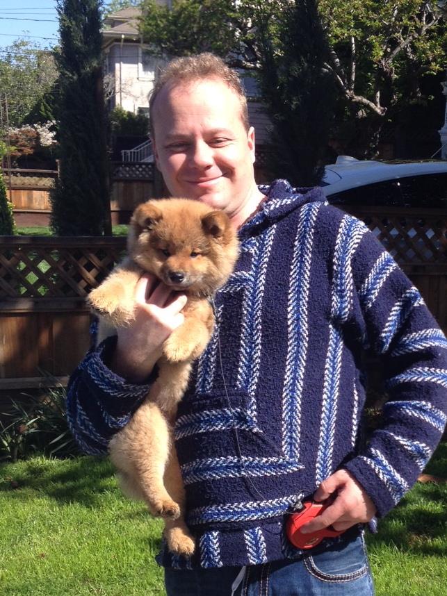 A man stands in a yard, joyfully holding a small, fluffy puppy on a sunny day.
