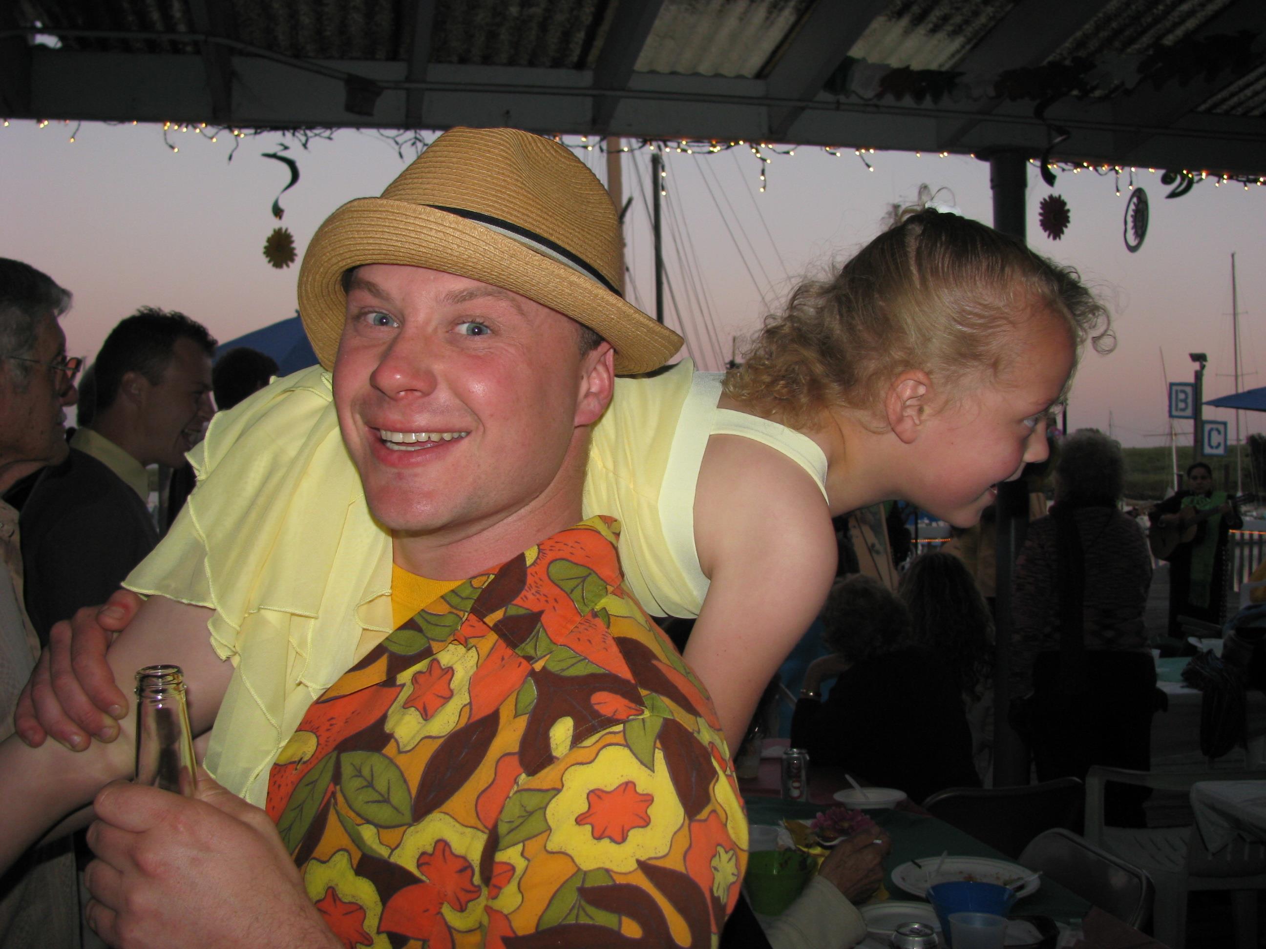 A man enjoys a fun moment with a child on his back amidst a festive beach event at dusk.