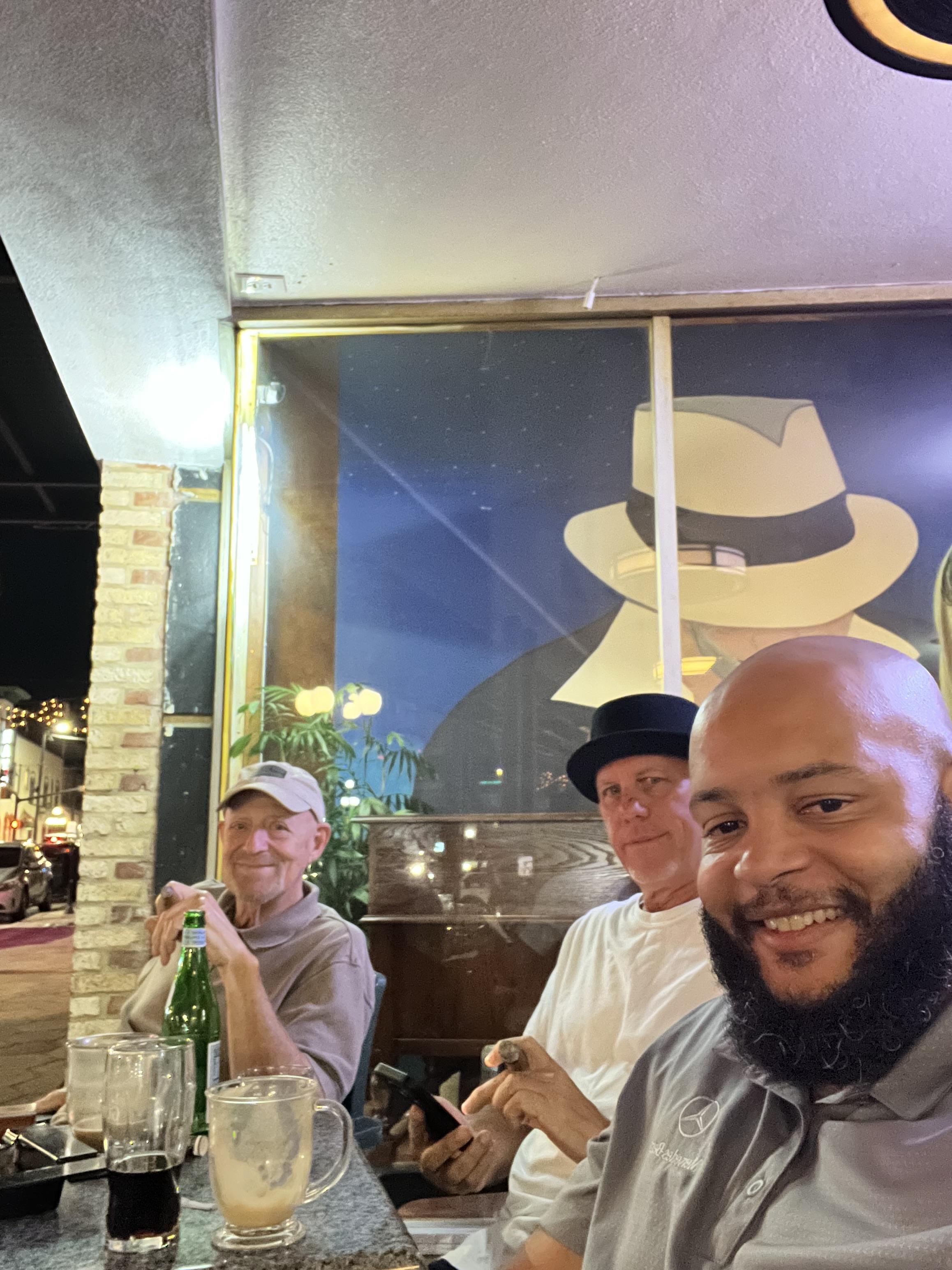 Three men sit together at a cafe table, sharing drinks and laughter on a lively evening.