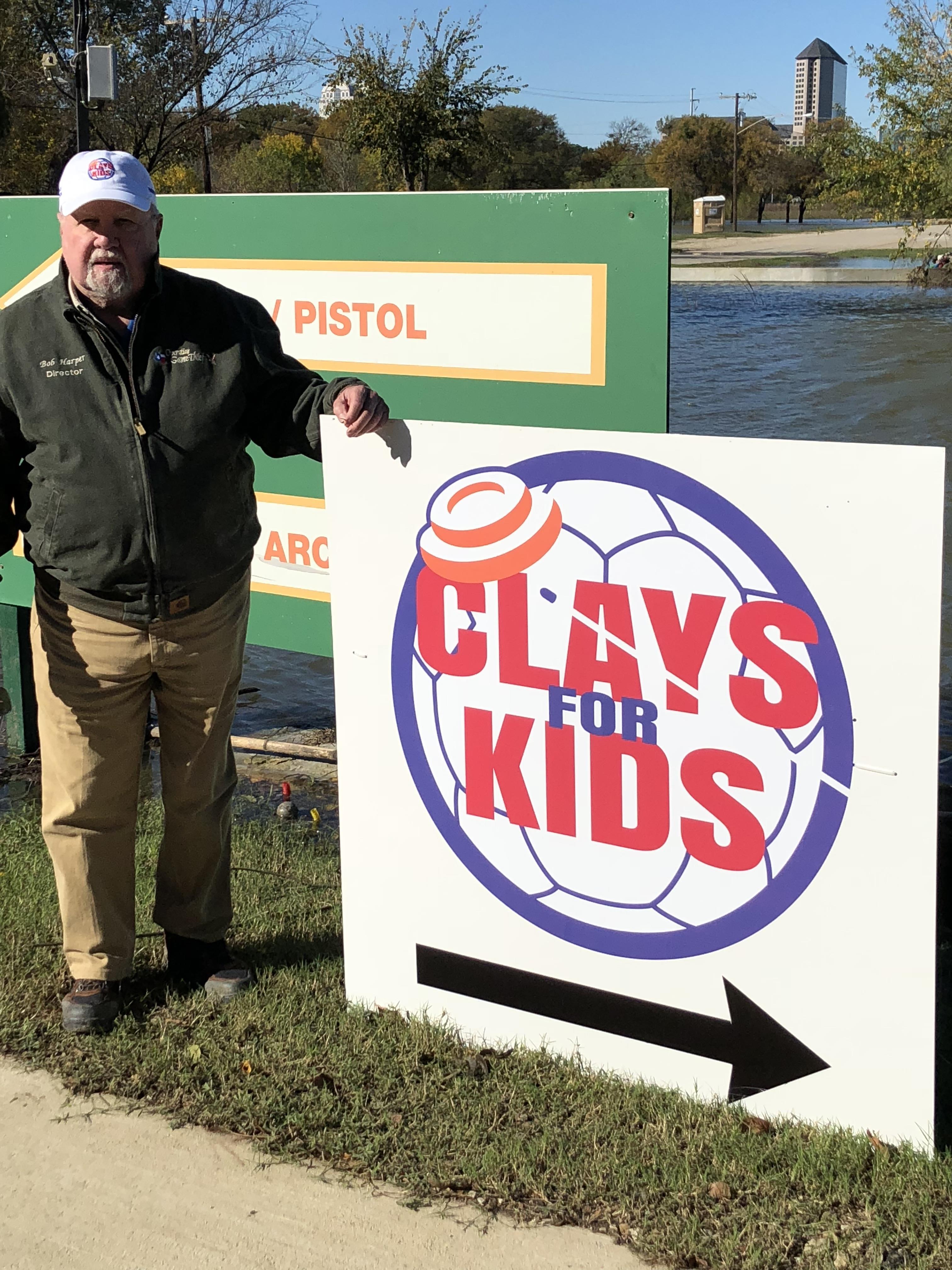 Person poses by a Clays for Kids sign by the water under clear blue skies in the park.
